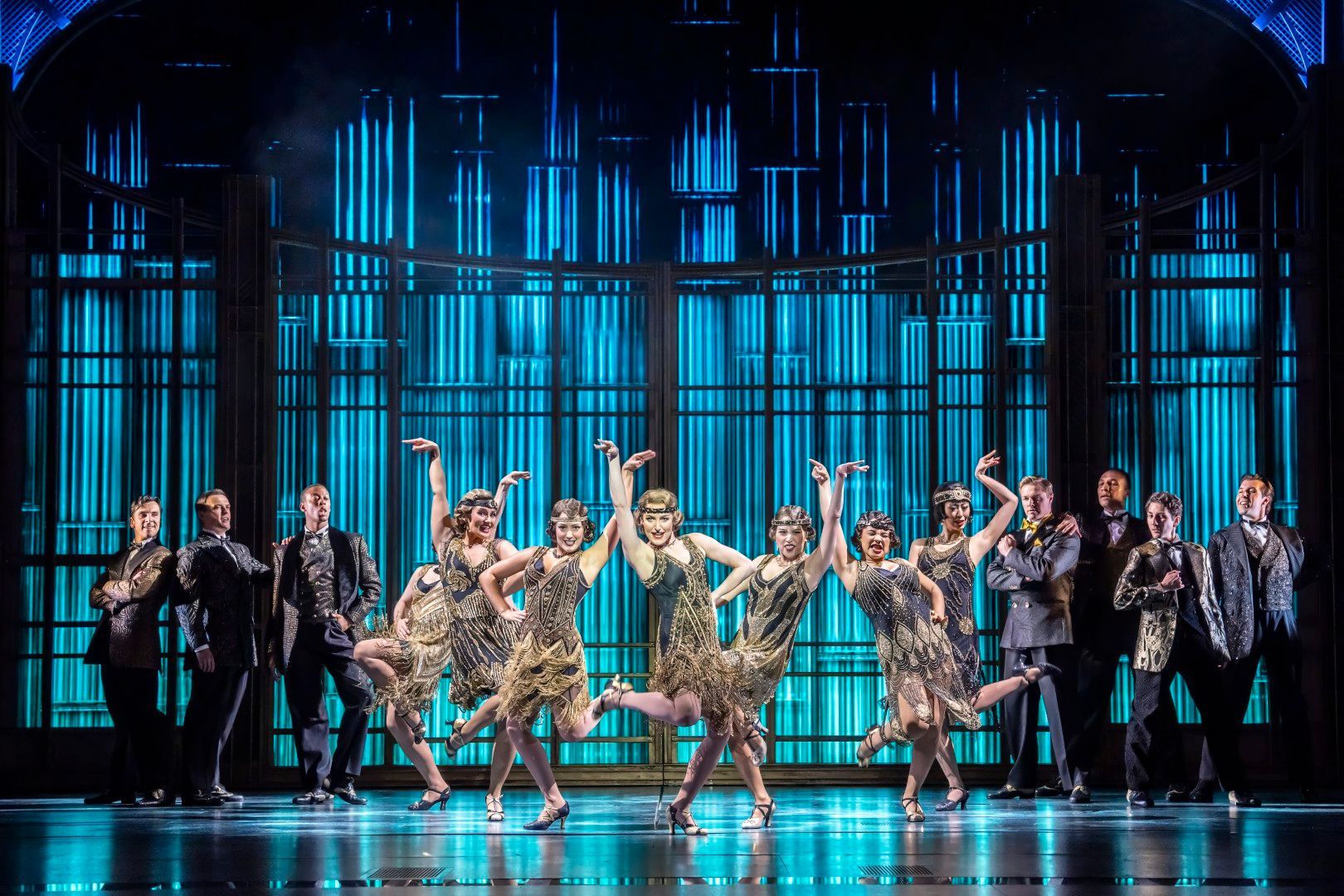 A wide shot of the cast in a dramatic tableau during one of Gatsby’s party scenes, with dancers in 1920s costumes and bright, theatrical lighting illuminating the stage.
