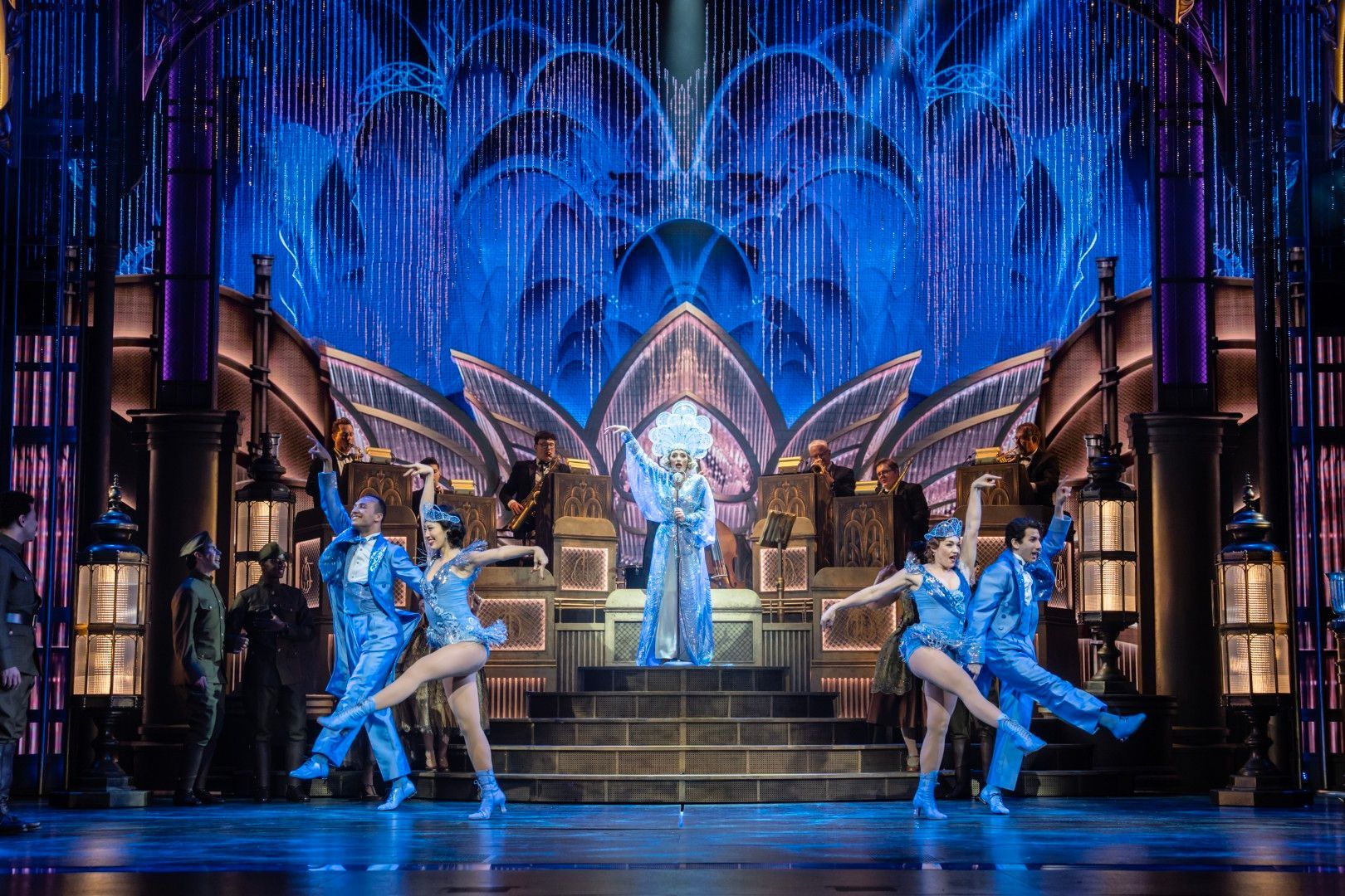 Dancers in sparkling blue 1920s costumes perform in front of a jazz band and shimmering art deco backdrop.