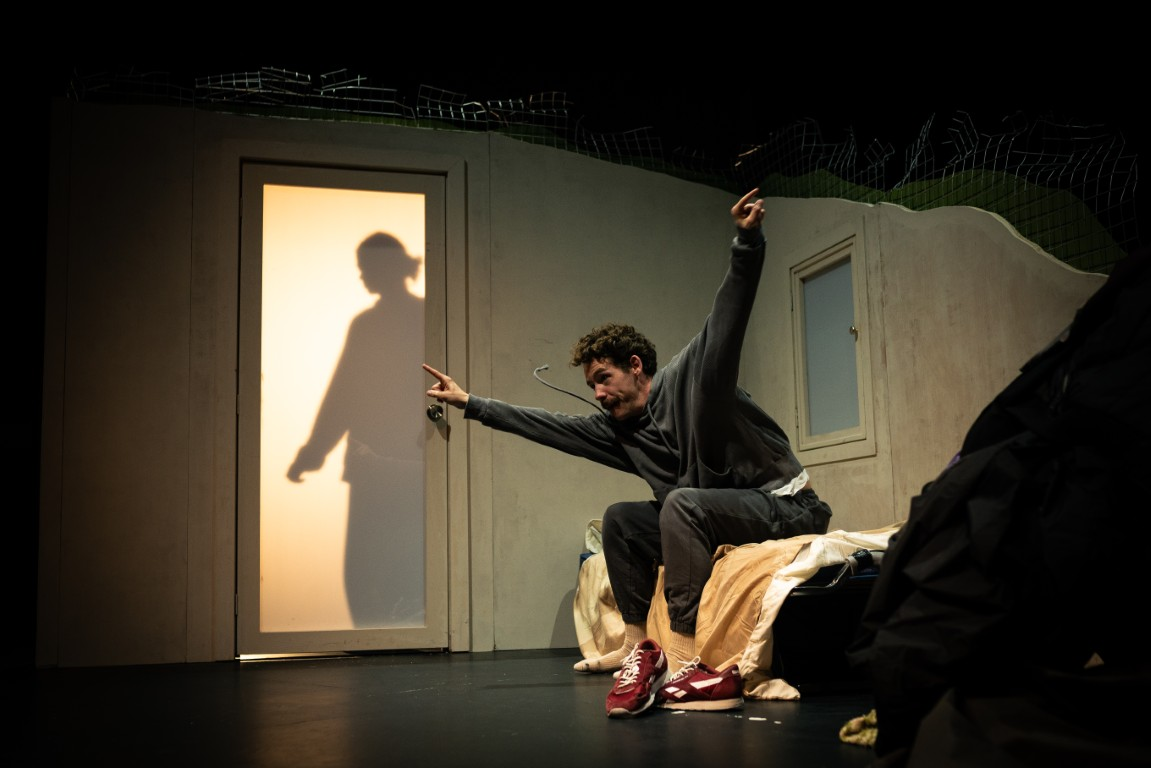 A man sits on a messy bed, pointing fingers in a celebratory gesture. Behind him, a shadowy silhouette looms on the door.