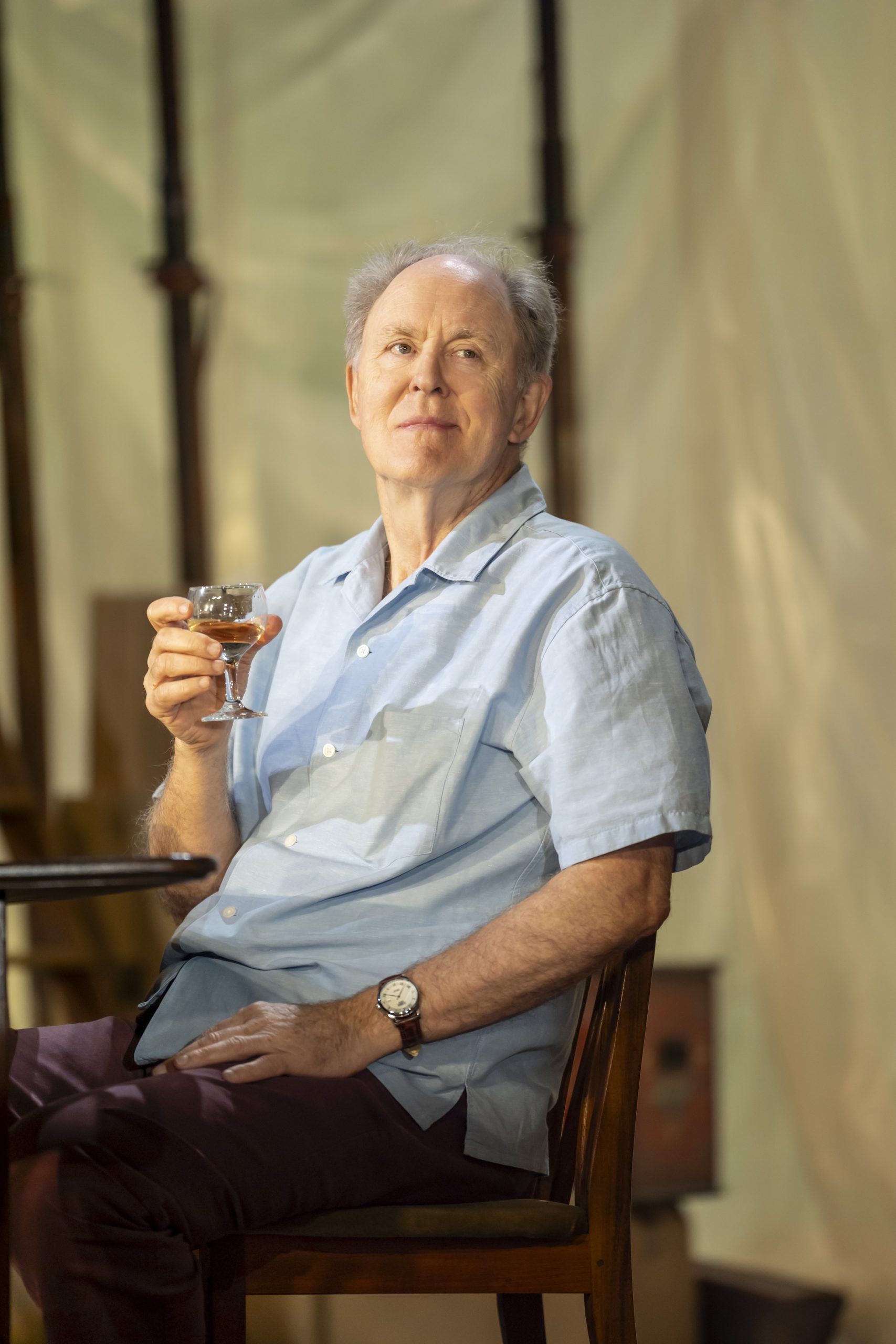 John Lithgow sits with a slight smile, holding a glass of wine and looking off to the side in a moment of reflection.