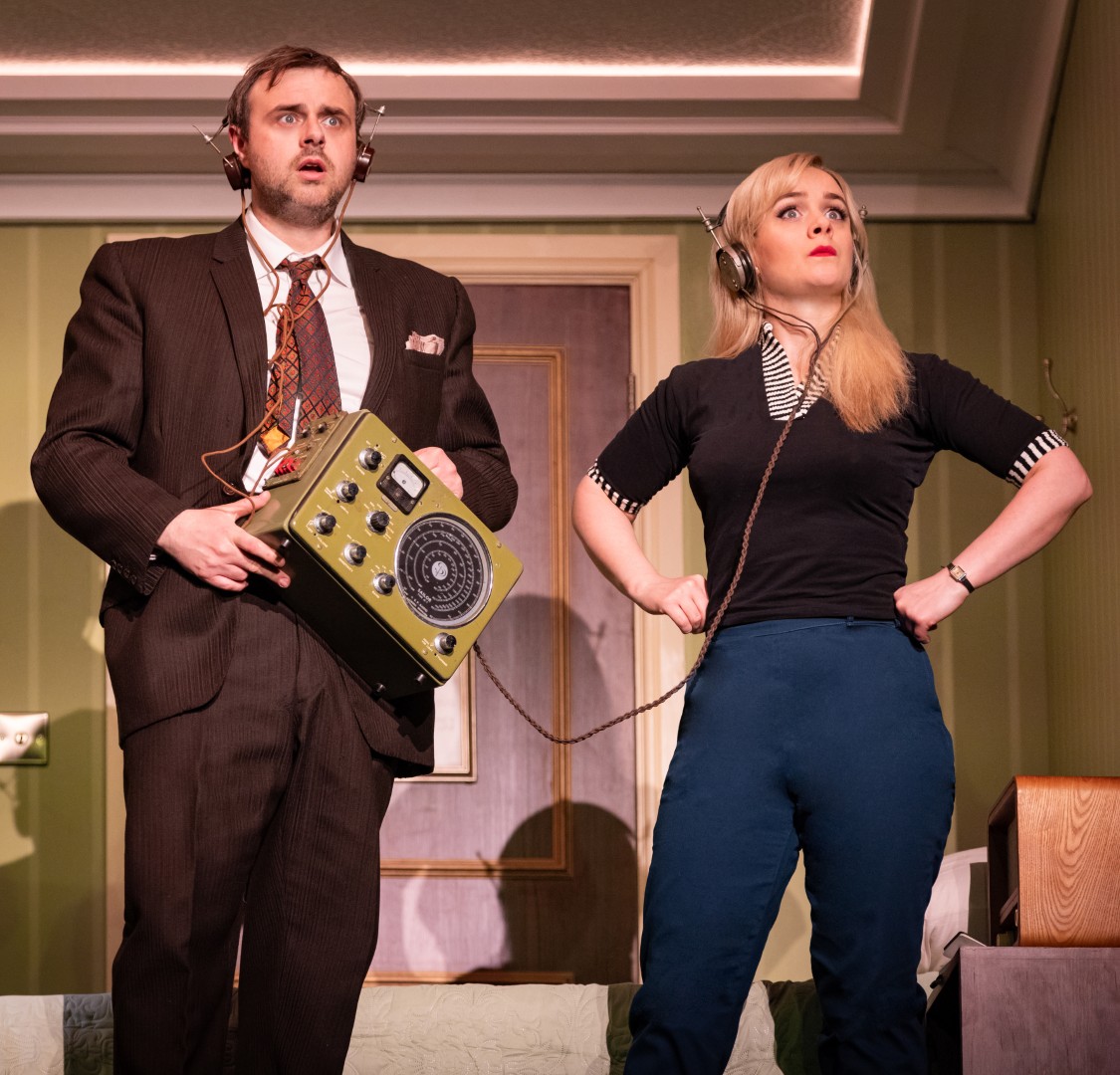 Chris Leask and Charlie Russell as Sergei and Elena wear headphones and hold spy equipment in a green hotel room