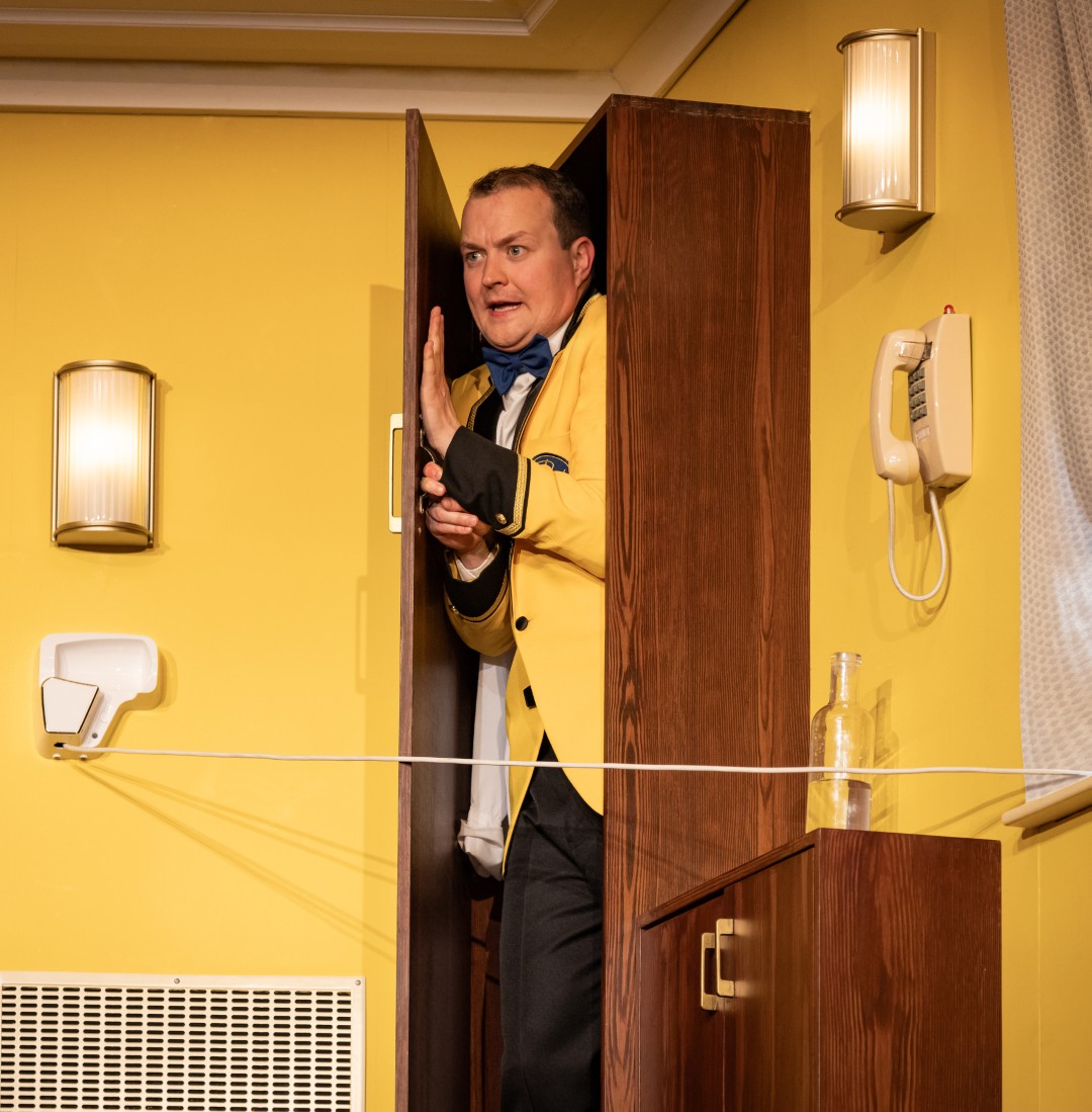 Greg Tannahill peeks nervously out from a wardrobe in a bright yellow hotel room, dressed in a bellboy uniform