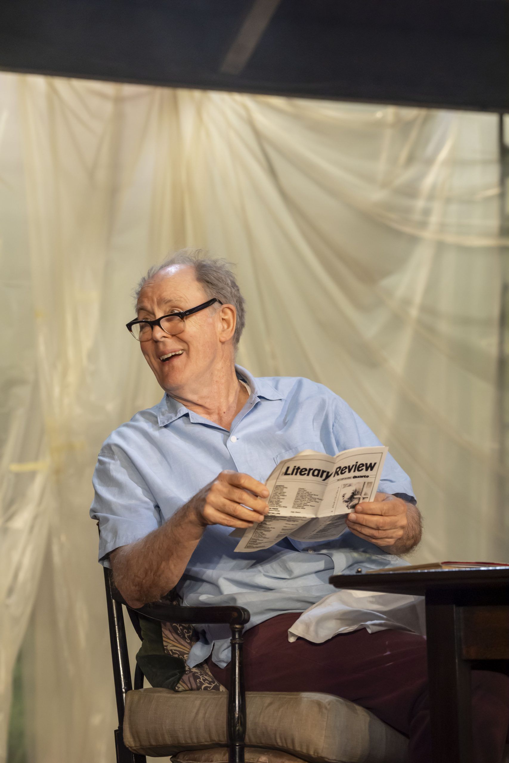 John Lithgow smiles while holding up a folded copy of Literary Review, seated on stage with draped plastic sheeting in the background.