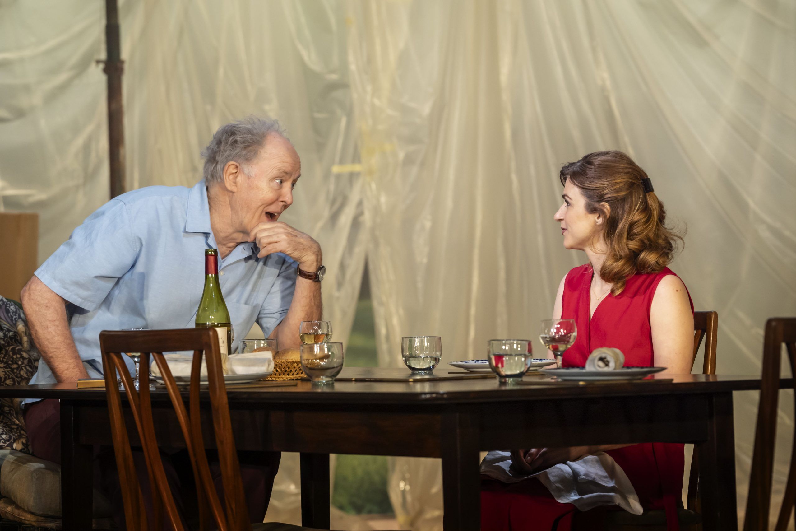 John Lithgow leans animatedly across the table toward Aya Cash, who listens attentively, seated opposite him in a red dress.