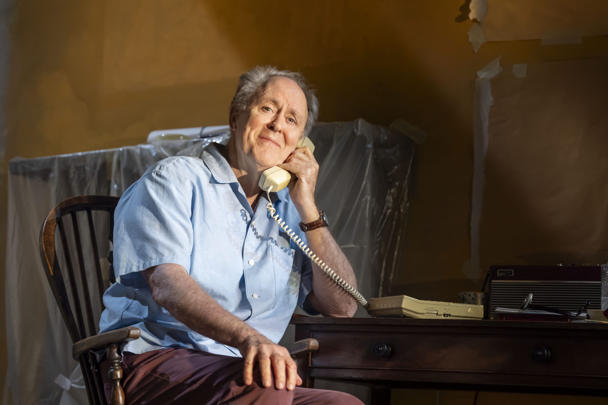 John Lithgow sits at a desk, smiling gently while holding a cream-coloured landline phone to his ear.