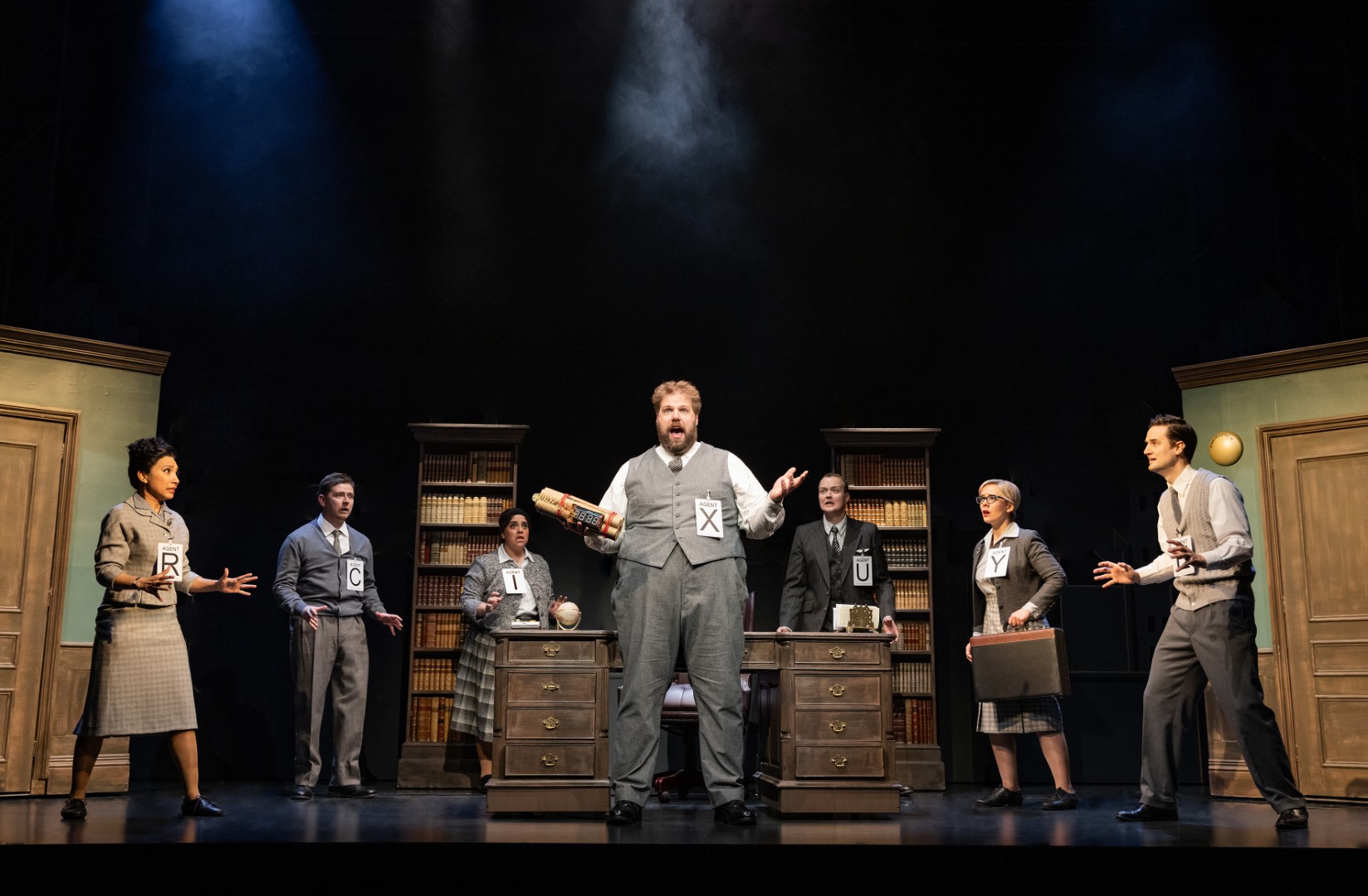Henry Lewis stands centre stage holding a bundle of TNT, surrounded by six other agents each labelled with a different letter