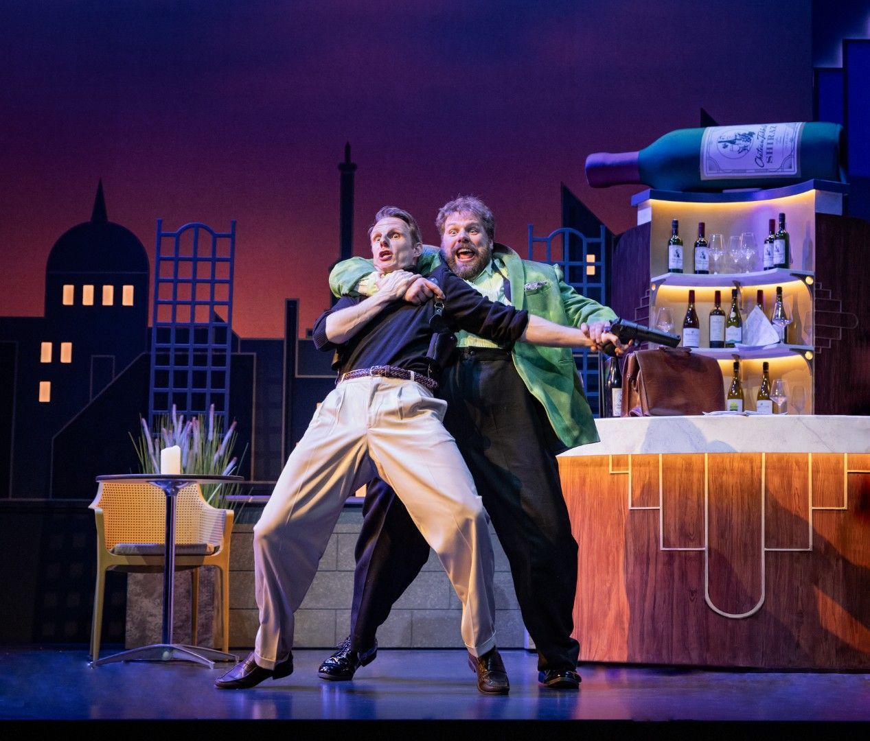 Henry Lewis grabs Dave Hearn in a headlock during a rooftop bar brawl, with a skyline backdrop and a giant bottle of Shiraz poised above the bar.