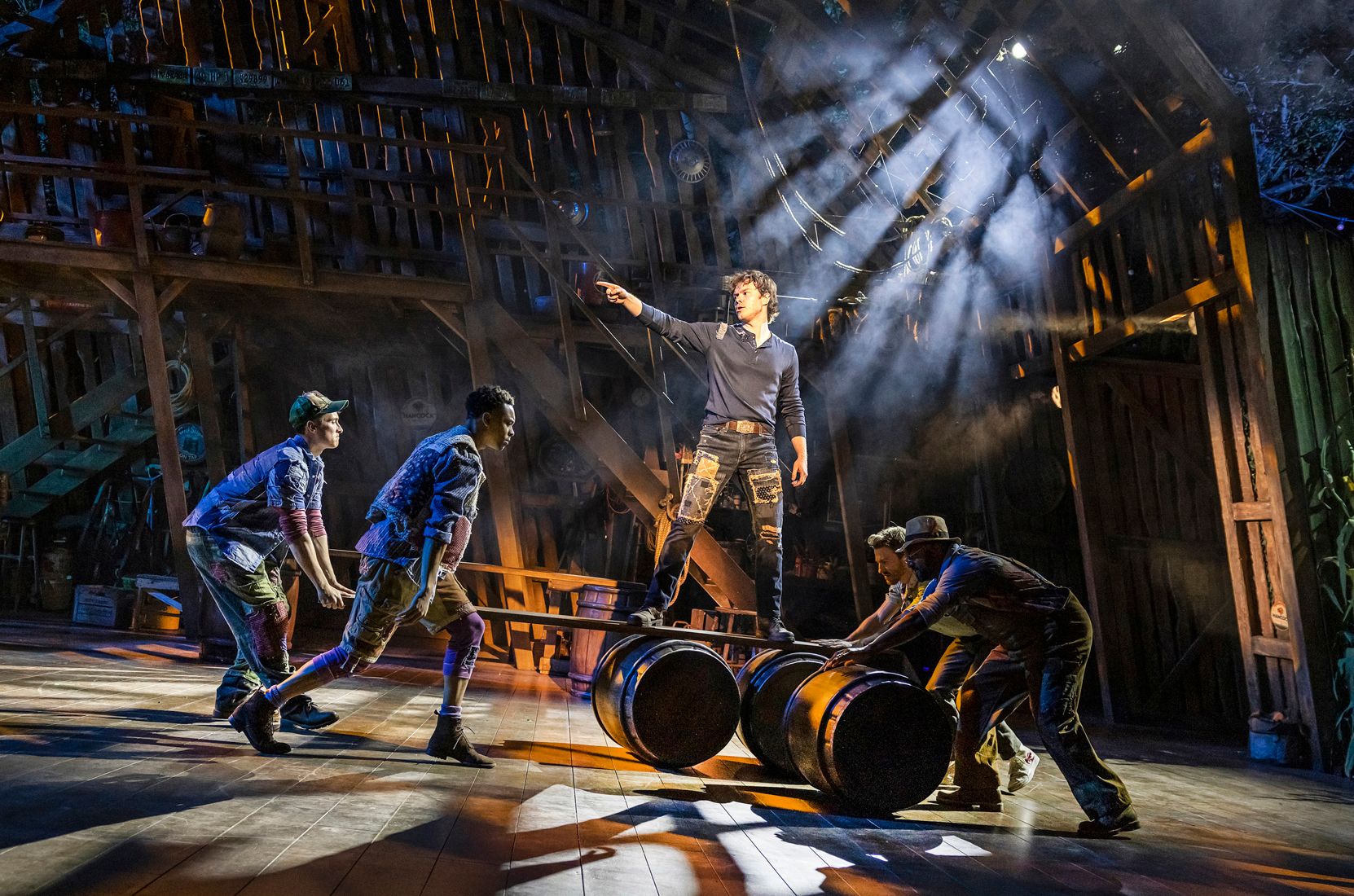 Ben Joyce as Beau balances heroically on barrels in a dramatic shaft of light, pointing forward, as four ensemble members support the barrels beneath him in a choreographed sequence.