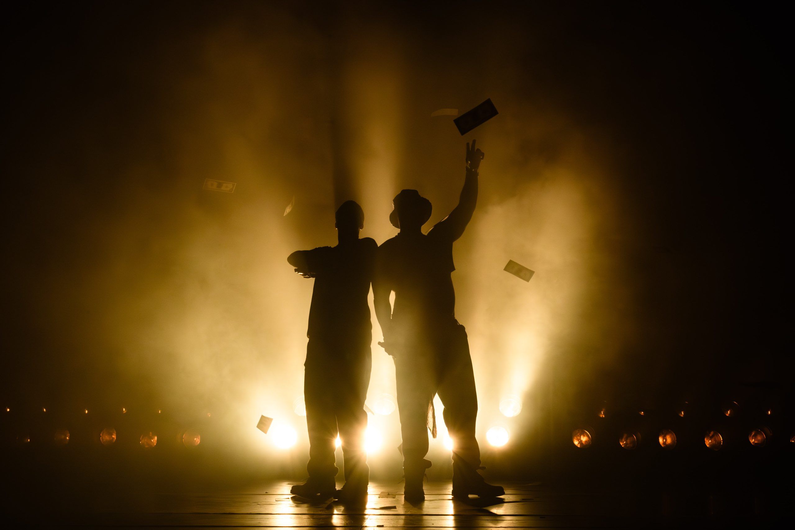 Silhouettes of Clifford Samuel and Daniel Ward standing in dramatic backlight with dollar bills floating through stage smoke.