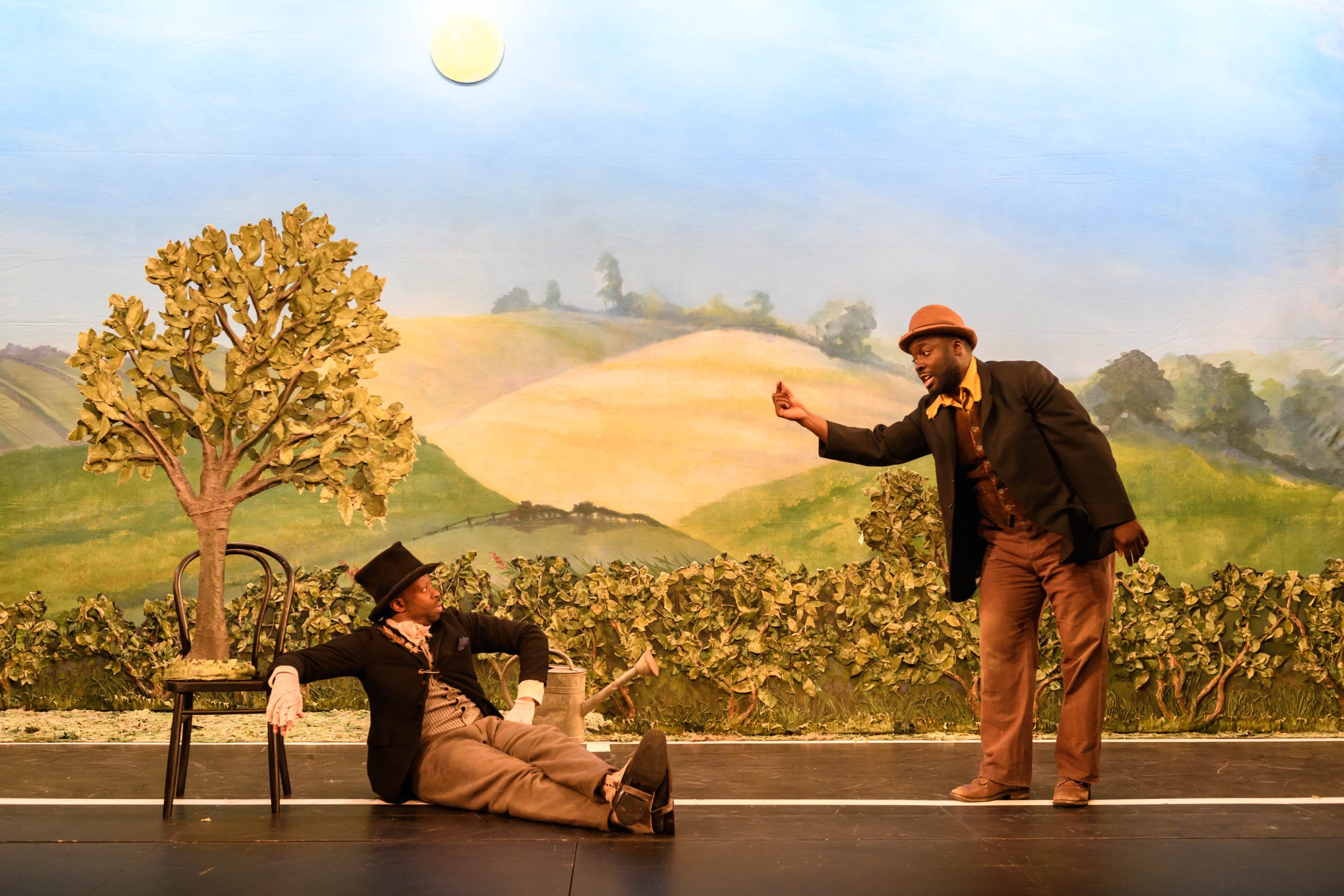 Daniel Ward offers a quarter to Clifford Samuel, who is reclining beneath a fake tree, in a stylised rural setting.