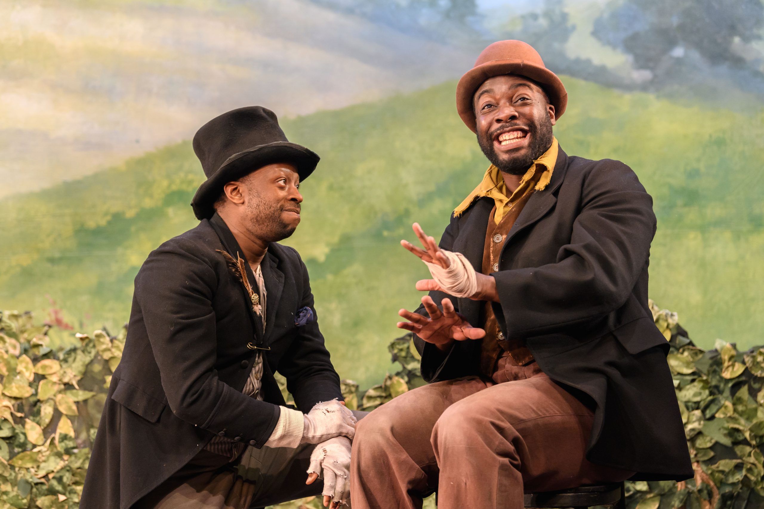 Clifford Samuel and Daniel Ward in early minstrel costumes, sitting in front of a fake countryside set, mid-dialogue.