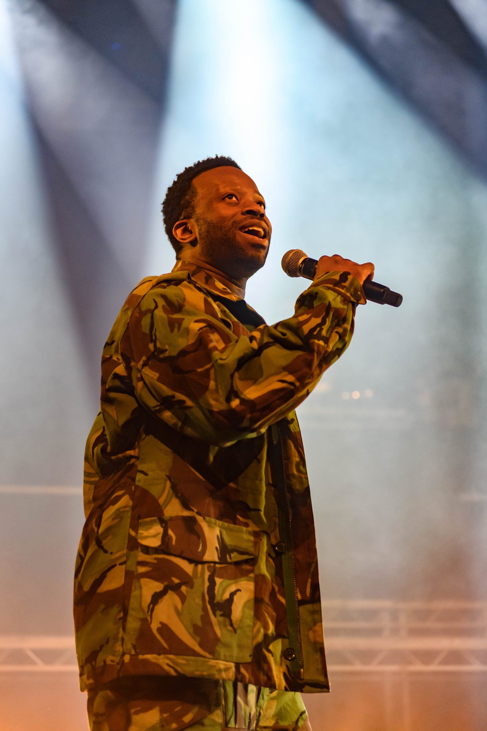 Clifford Samuel in camouflage jacket holding a mic, looking up into stage lights with an expressive face.