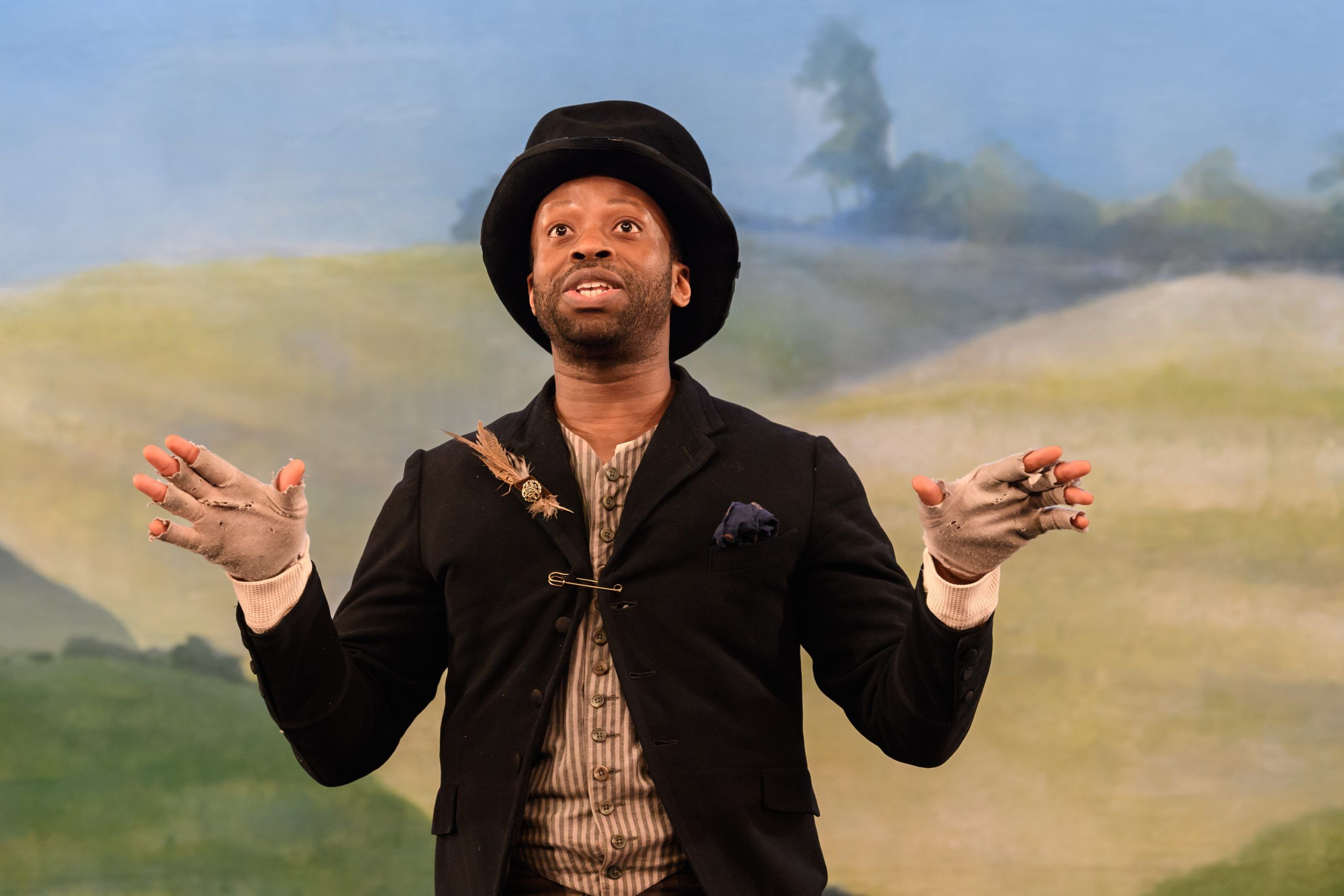 Clifford Samuel in a top hat and tattered gloves, gesturing passionately in front of a painted countryside backdrop.