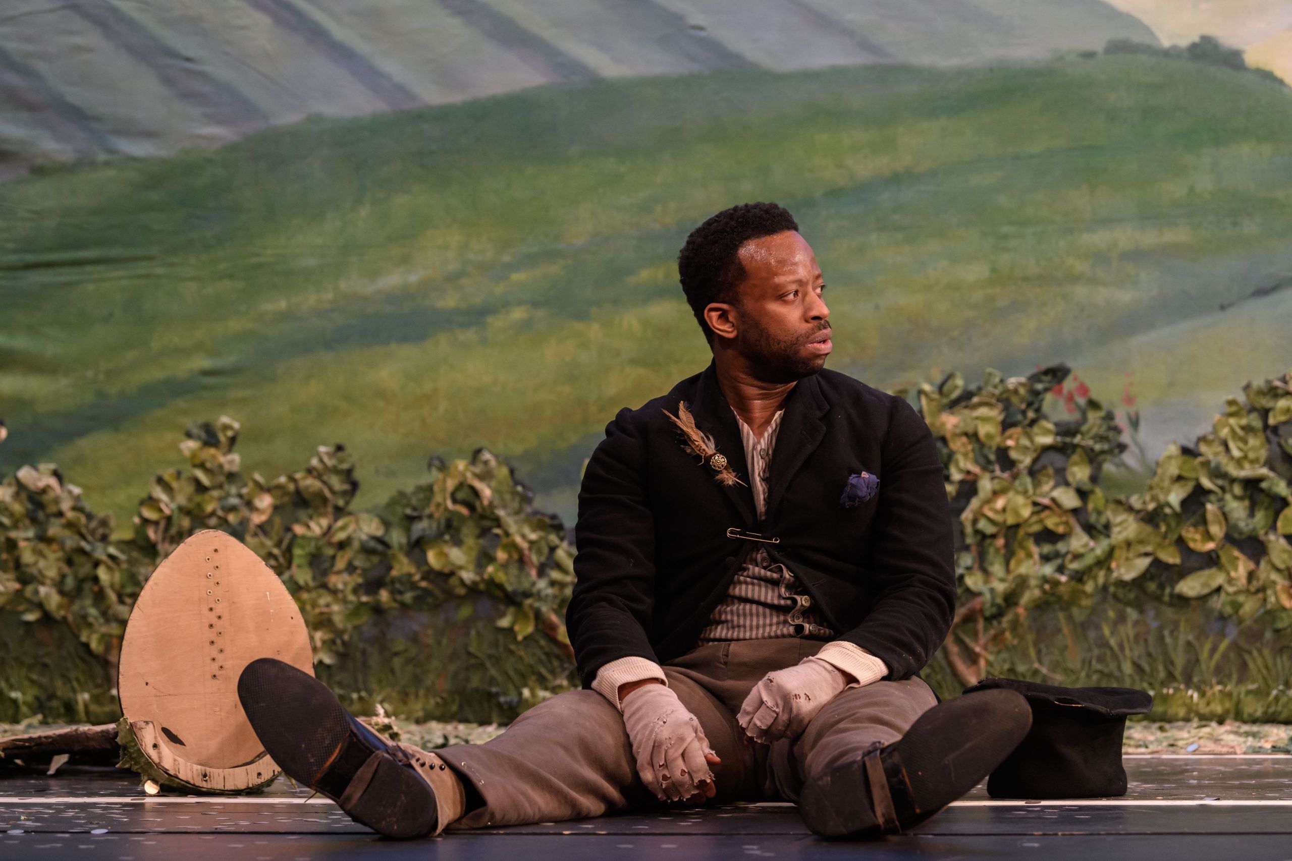 Clifford Samuel sitting on stage, legs stretched out, gazing sideways against the fake grass and painted hills.