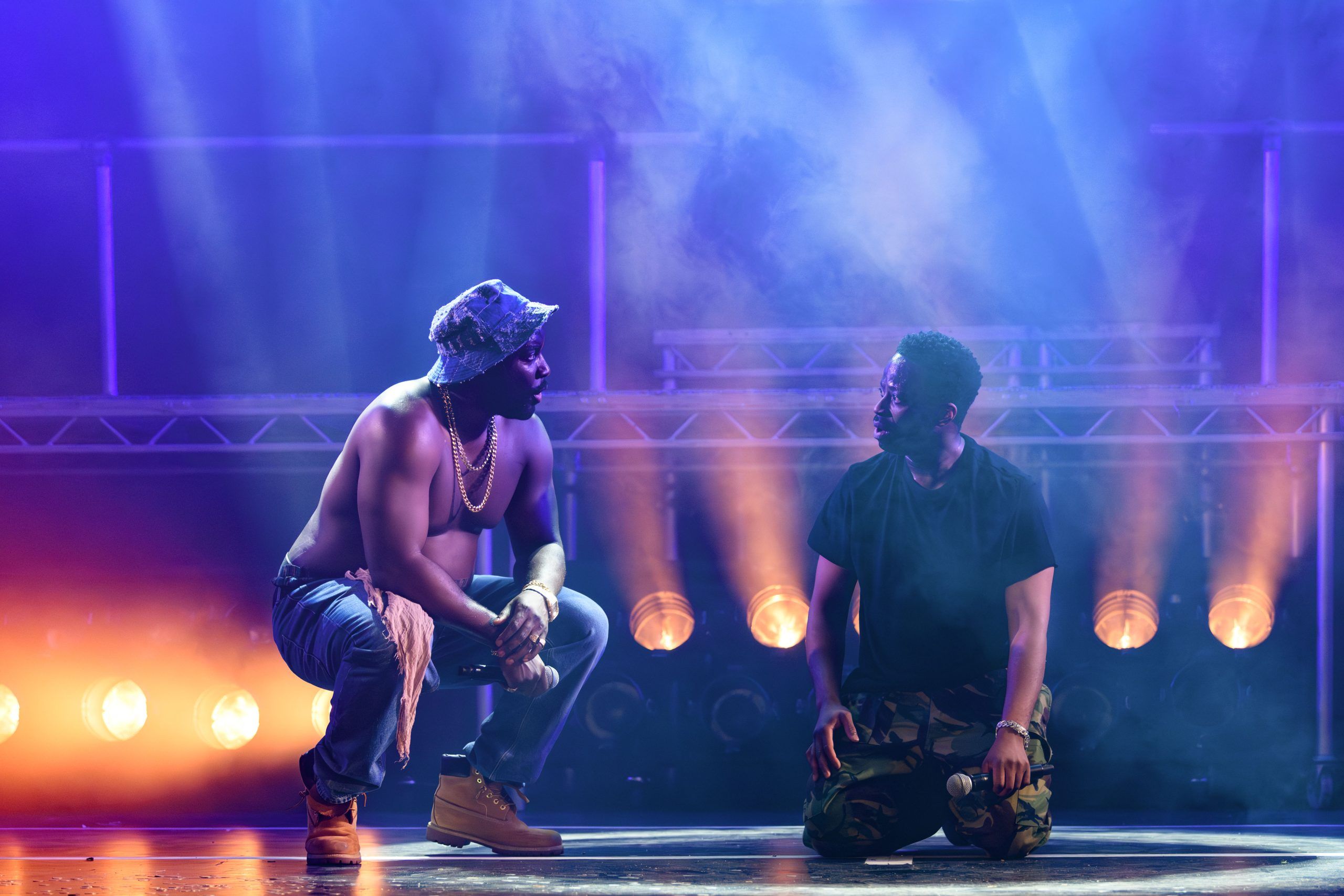 Daniel Ward and Clifford Samuel crouched face-to-face, caught in an intense moment, bathed in purple and orange light.