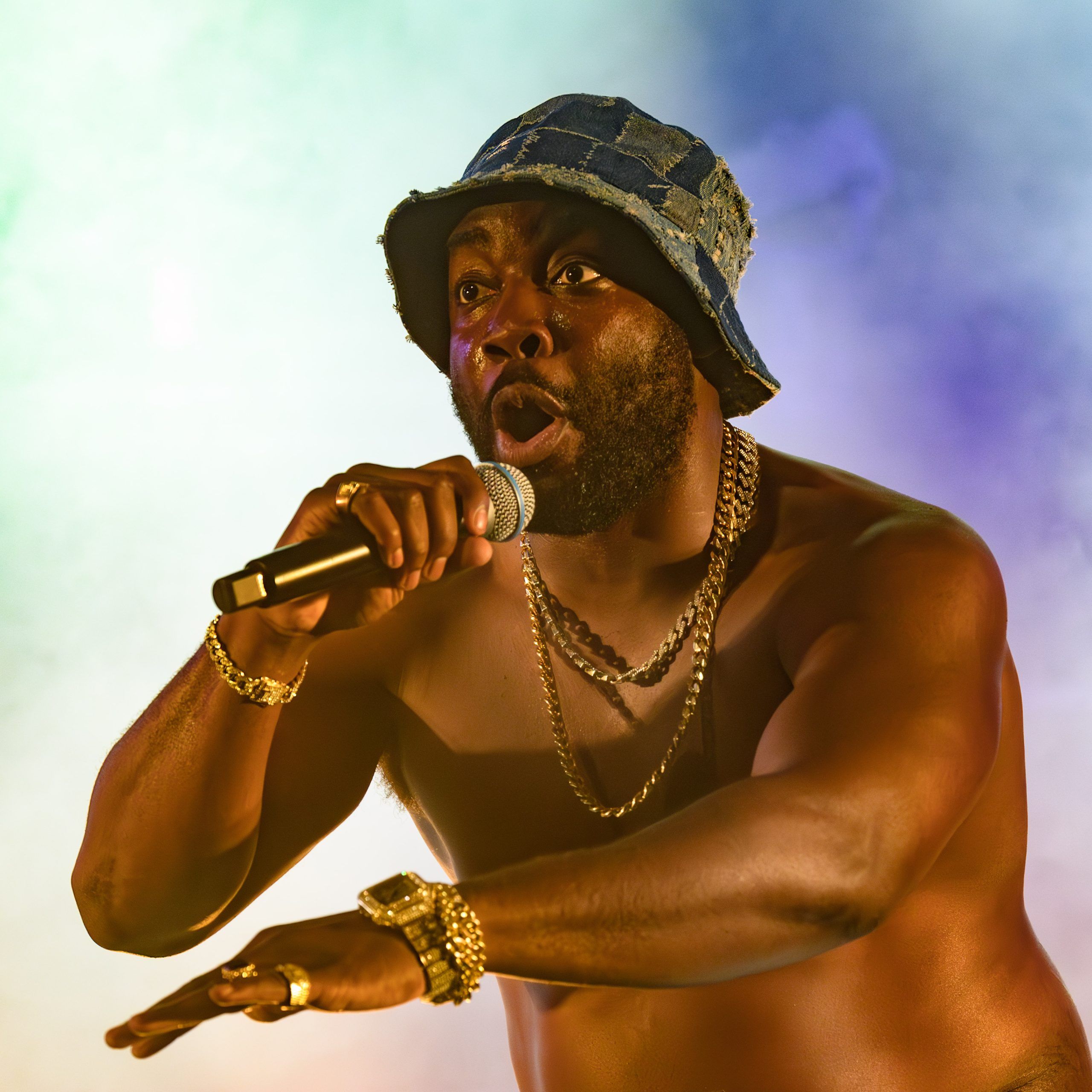 Daniel Ward shirtless and intense, mic in hand, mouth open mid-line, framed by colourful stage smoke.