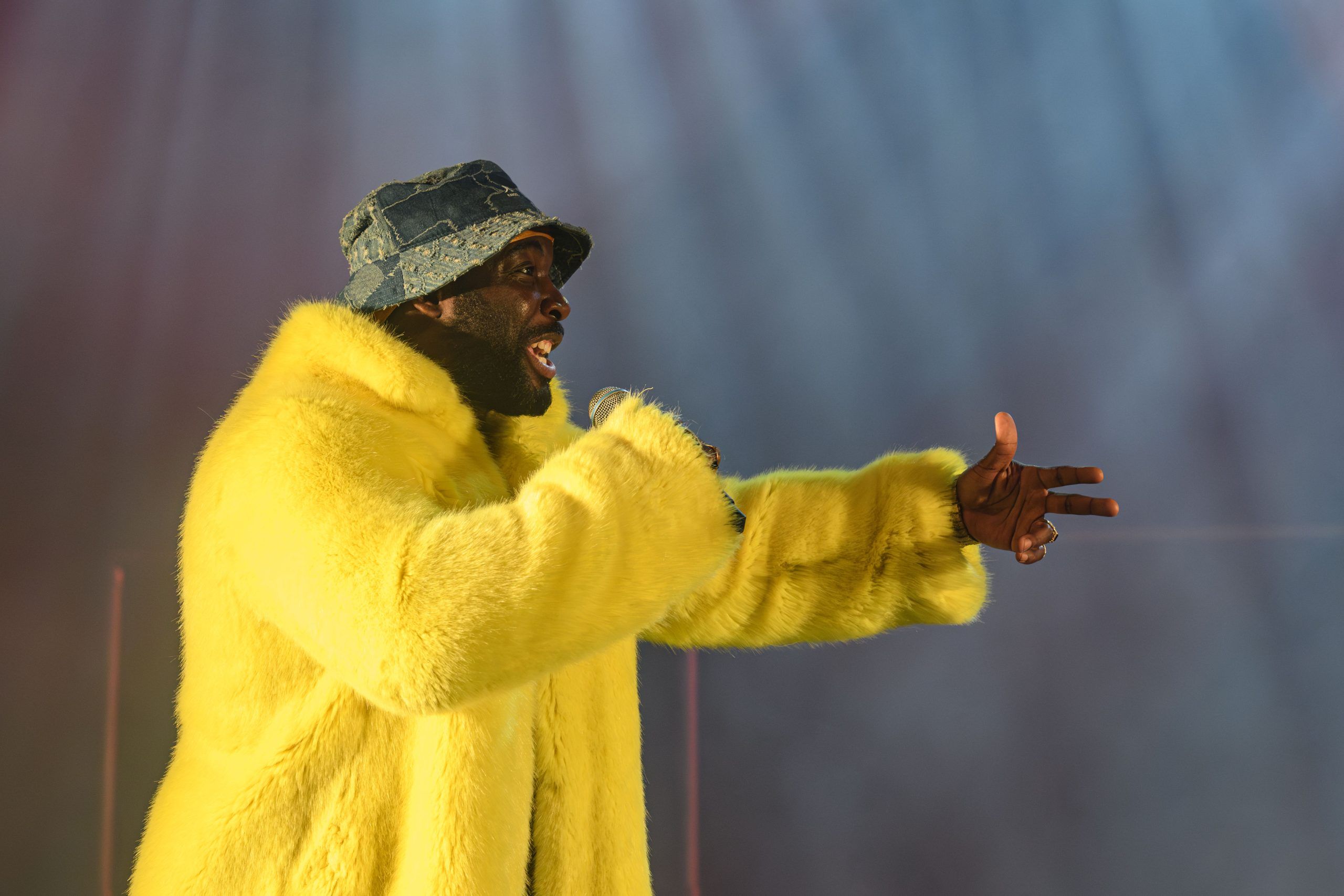 Daniel Ward in yellow fur and denim bucket hat, pointing and performing in a burst of dramatic stage light.