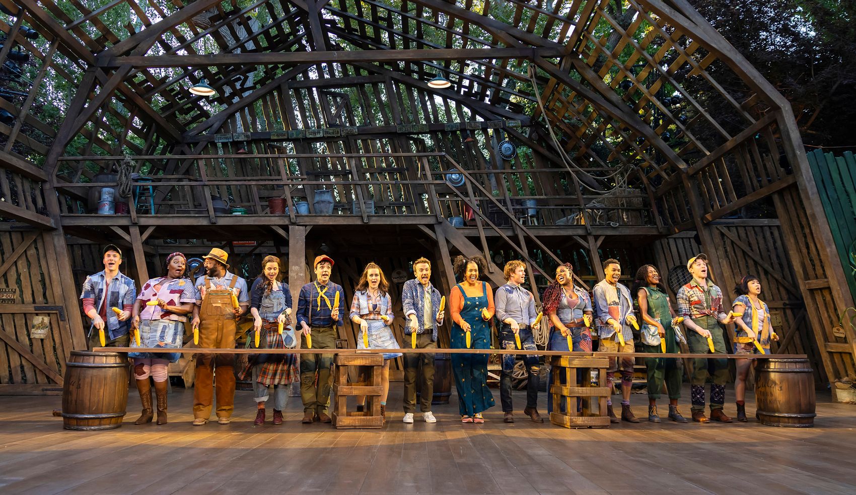 The full cast of Shucked lines up in a barn setting, each holding an ear of corn and singing in unison, dressed in colourful patchwork farmwear.
