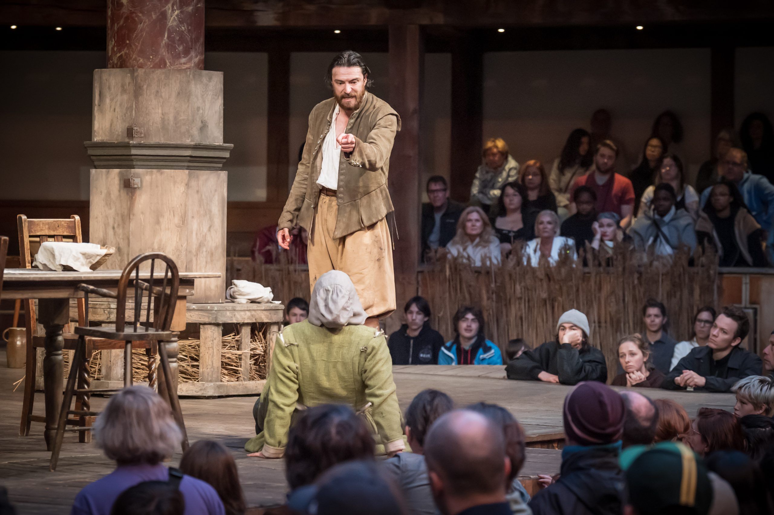 John Proctor, played by Gavin Drea, towers above a kneeling figure as he points with fury. The audience watches closely — part of the courtroom themselves.