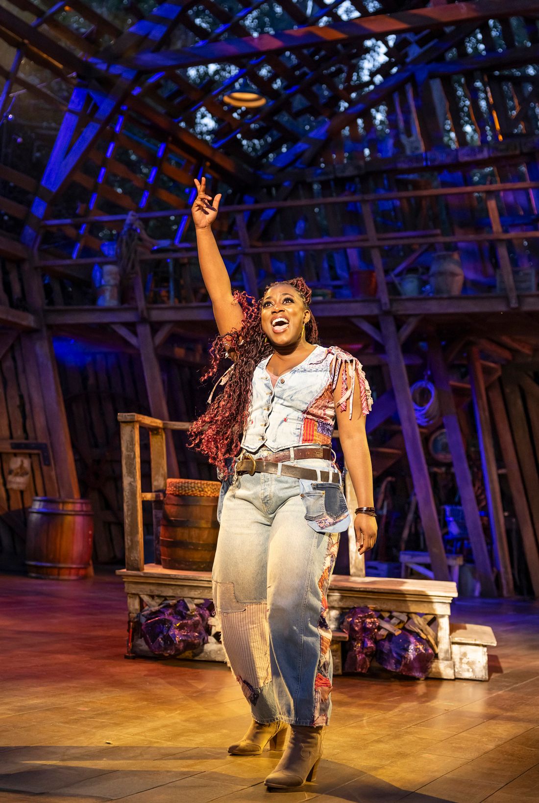 Georgina Onuorah as Lulu commands the stage with one arm raised, wearing patchwork jeans and a fitted denim vest, lit by warm amber stage lights.