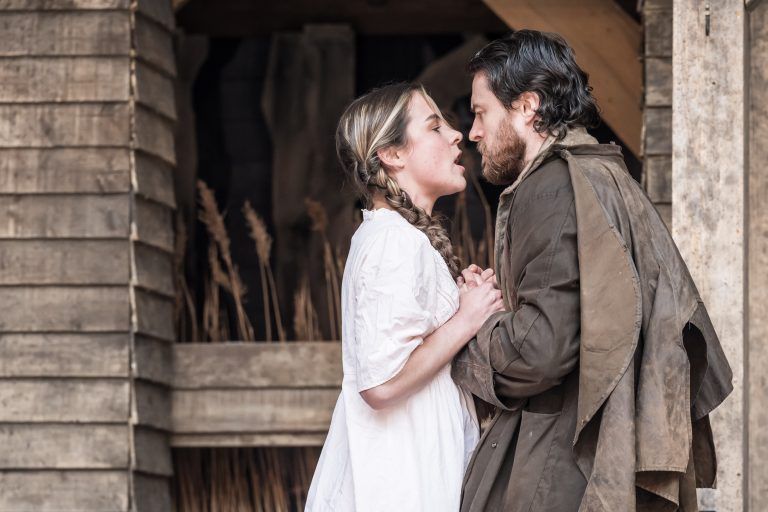 Abigail and Proctor face each other in close proximity — Hannah Saxby and Gavin Drea locked in a tense, breath-held moment, hands entwined, past and desire flaring.