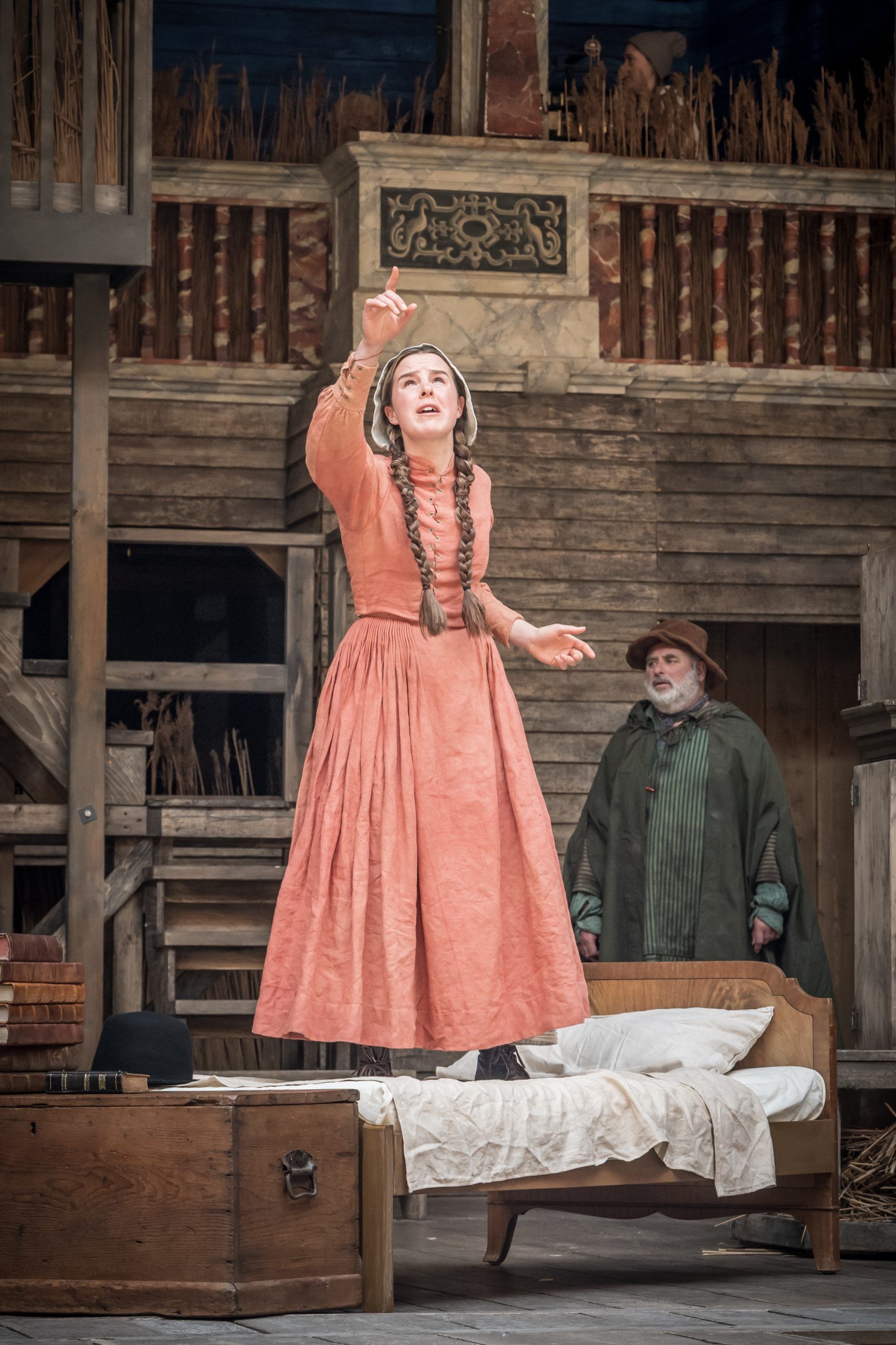 Abigail Williams, played by Hannah Saxby, stands atop a wooden bed, arm raised and finger pointing skyward in accusation, while chaos stirs behind her.