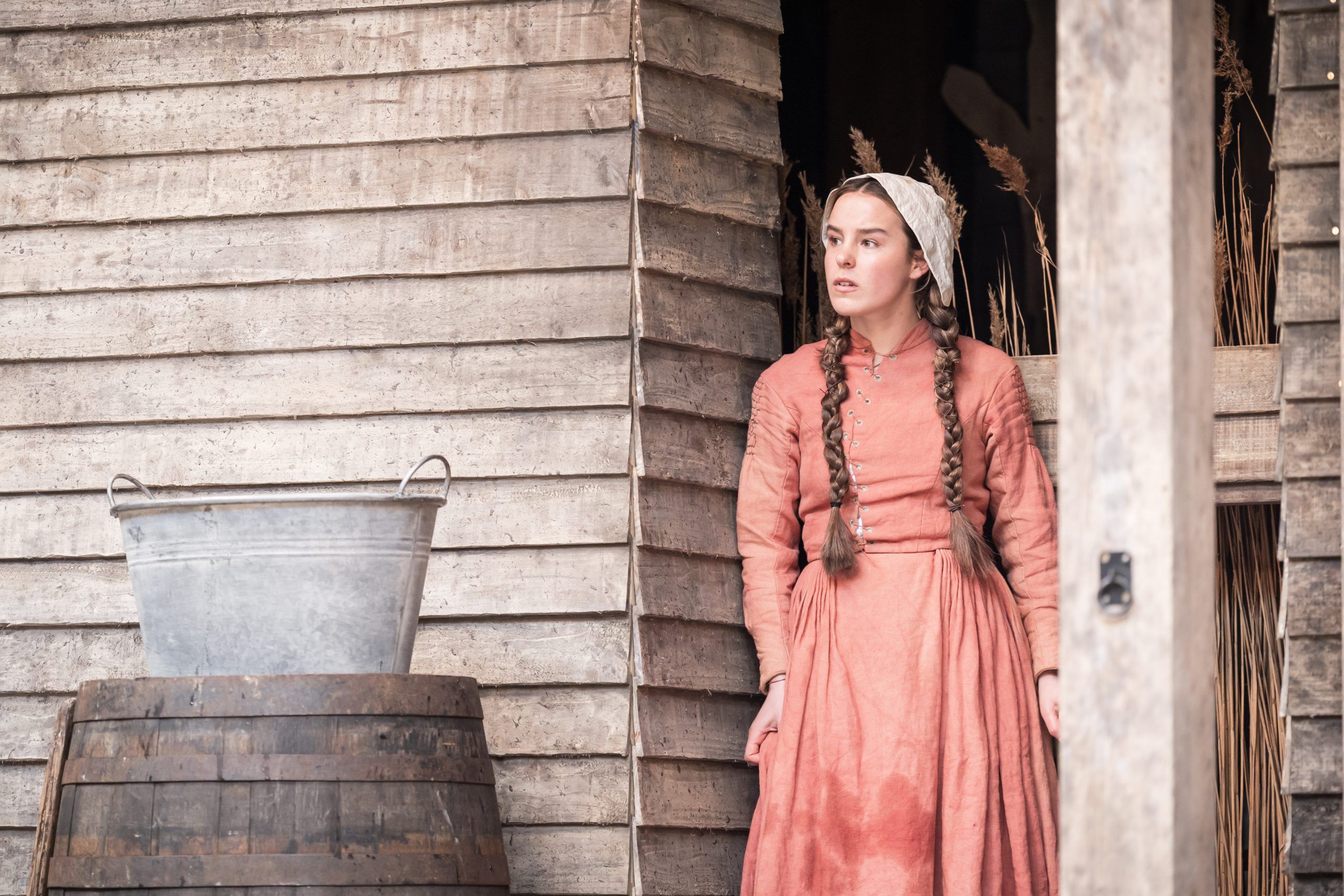 Hannah Saxby as Abigail Williams leans in the doorway of a weathered wooden structure, her long braids and bonnet framing a tense, watchful expression.