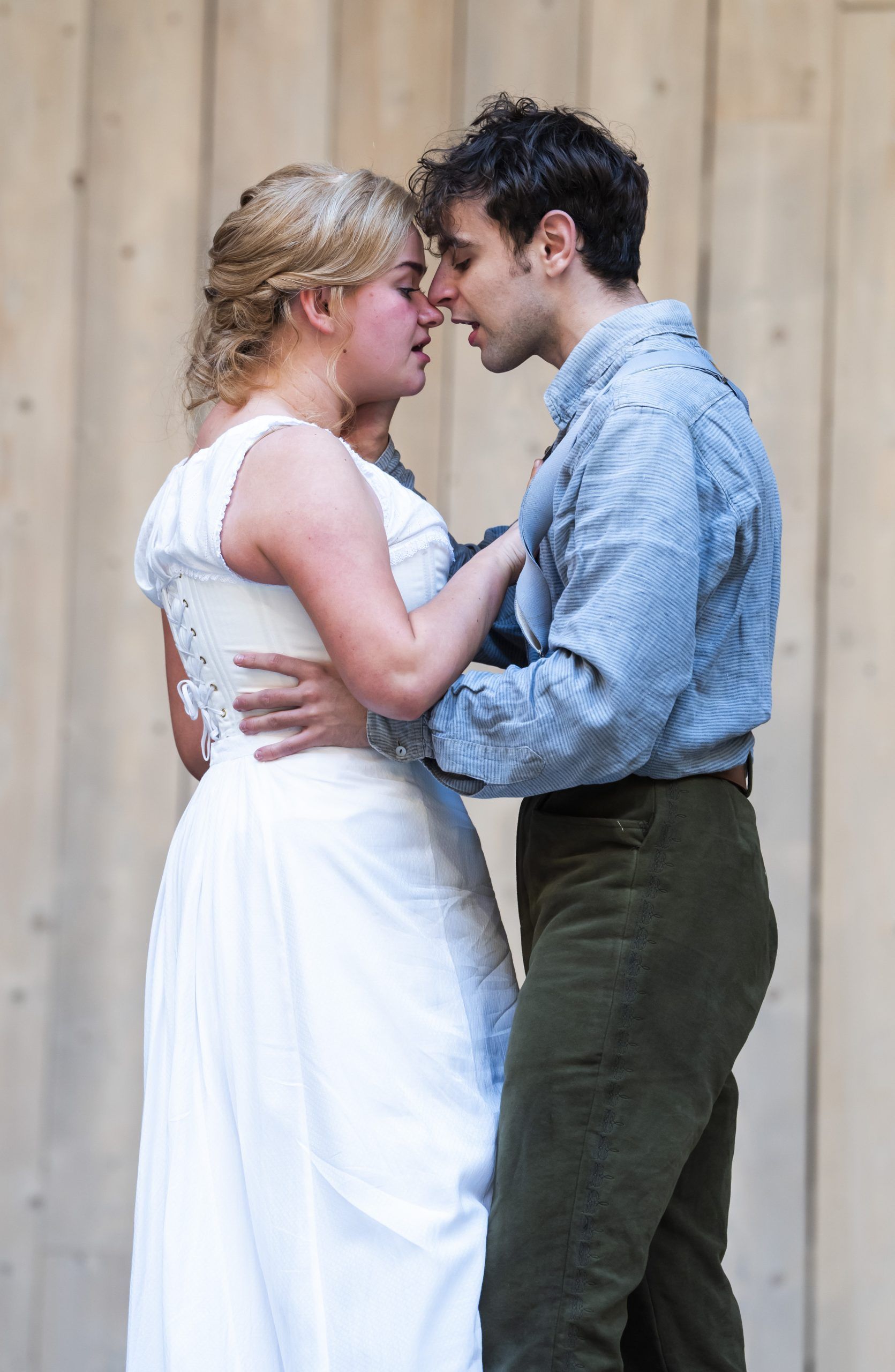 Lola Shalam and Rawaed Asde stand close together, foreheads almost touching in an intimate moment, both in soft focus against the wooden backdrop of the Globe stage.