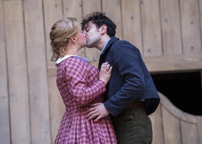 Lola Shalam and Rawaed Asde share a tender kiss in front of the wooden saloon doors, capturing a moment of intimacy on the Globe stage.