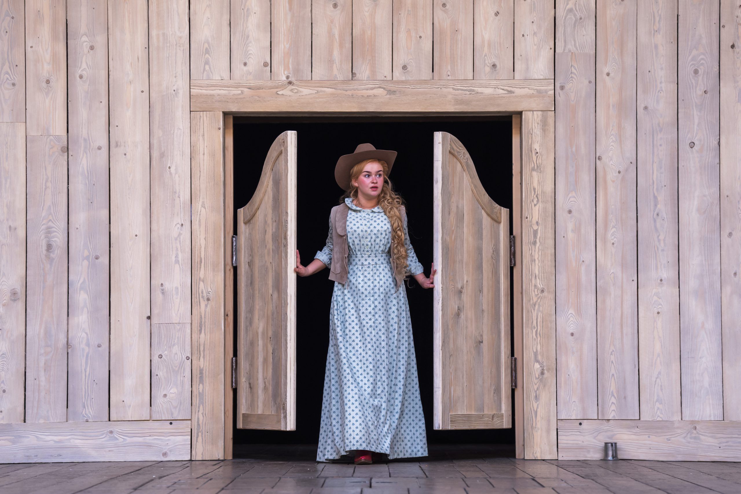 Lola Shalam steps through swinging saloon doors in a long floral dress and cowboy hat, framed by the wooden set of the Globe stage.