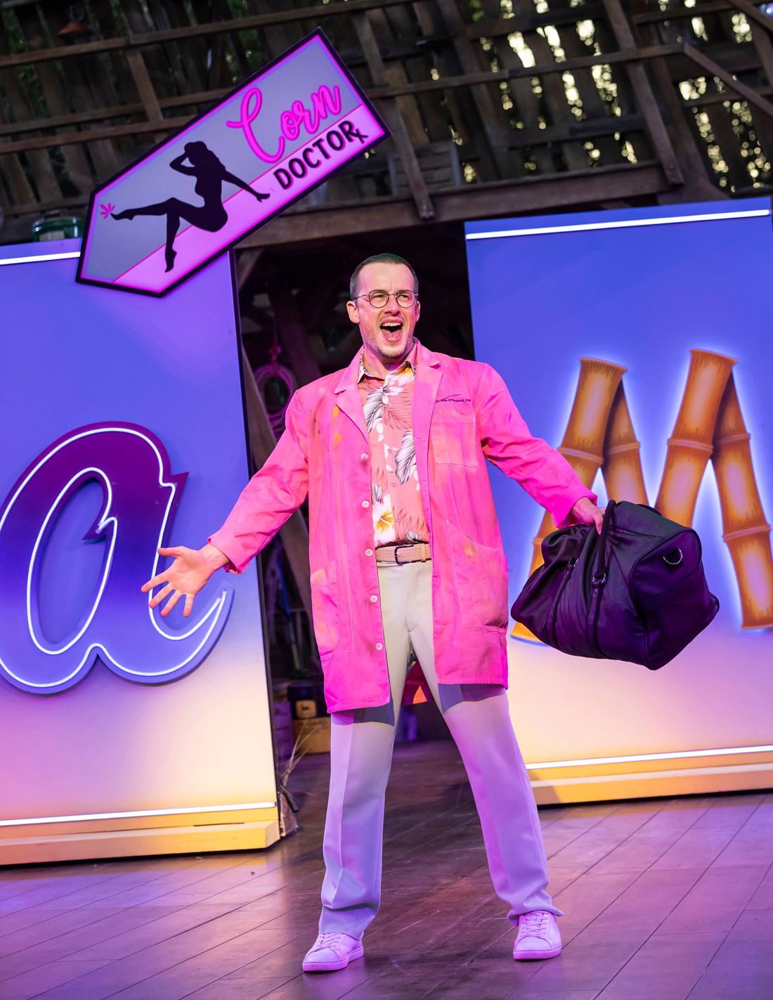 Matthew Seadon-Young as Gordy stands beaming under a neon pink “Corn Doctor” sign, wearing a bright pink coat, floral shirt, and pale trousers, holding a black duffel bag mid-song.