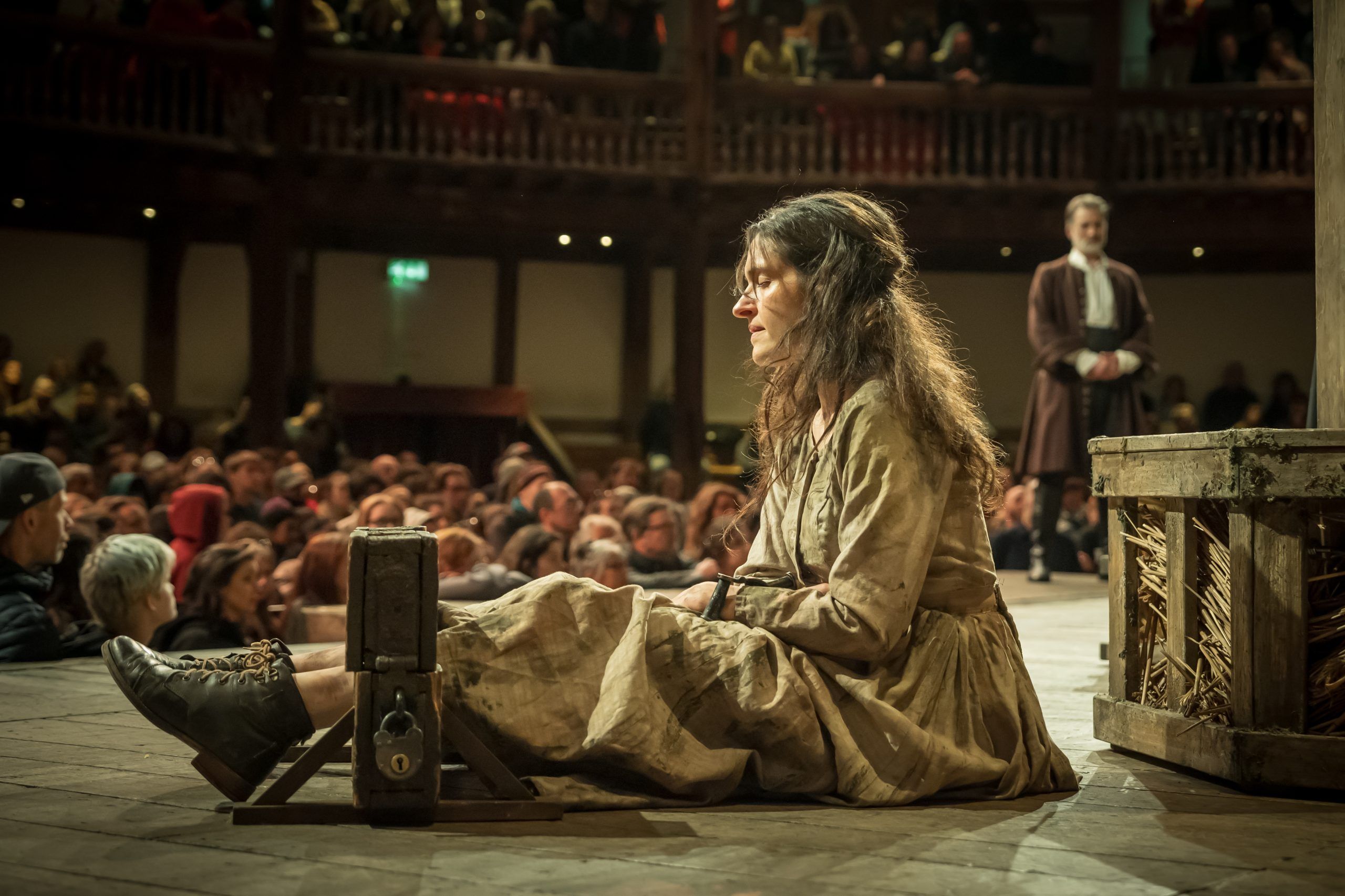 Phoebe Pryce sits in worn beige dress and shackles, her expression heavy with sorrow. Behind her, figures watch from the crowd and the stage alike — judgment hanging in the air.