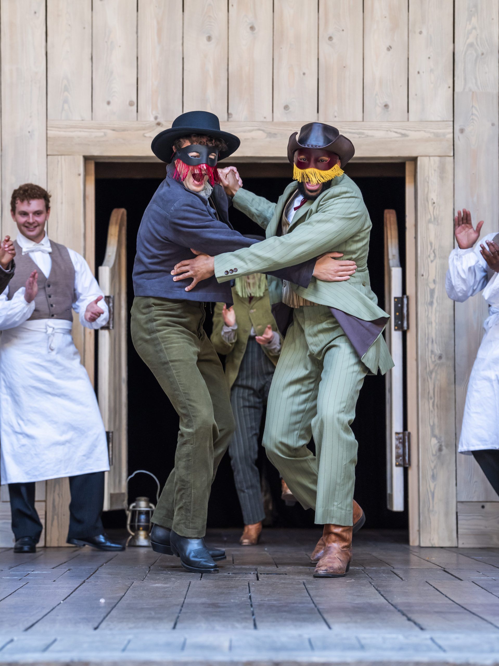 Rawaed Asde as Romeo and Michael Elcock as Mercutio dance joyfully in fringed masquerade masks, mid-celebration, flanked by clapping cast members outside the swinging saloon doors.