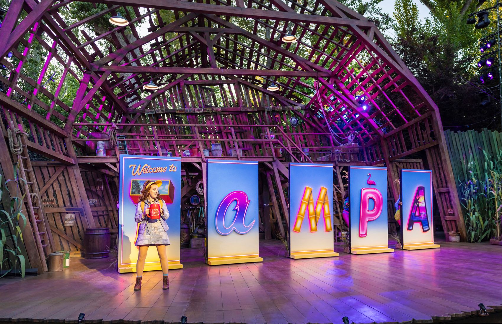 Sophie McShera stands before five colourful light-up panels spelling “TAMPA,” wearing a sunhat and holding a red Big Gulp mug in a bright, playful Florida scene.