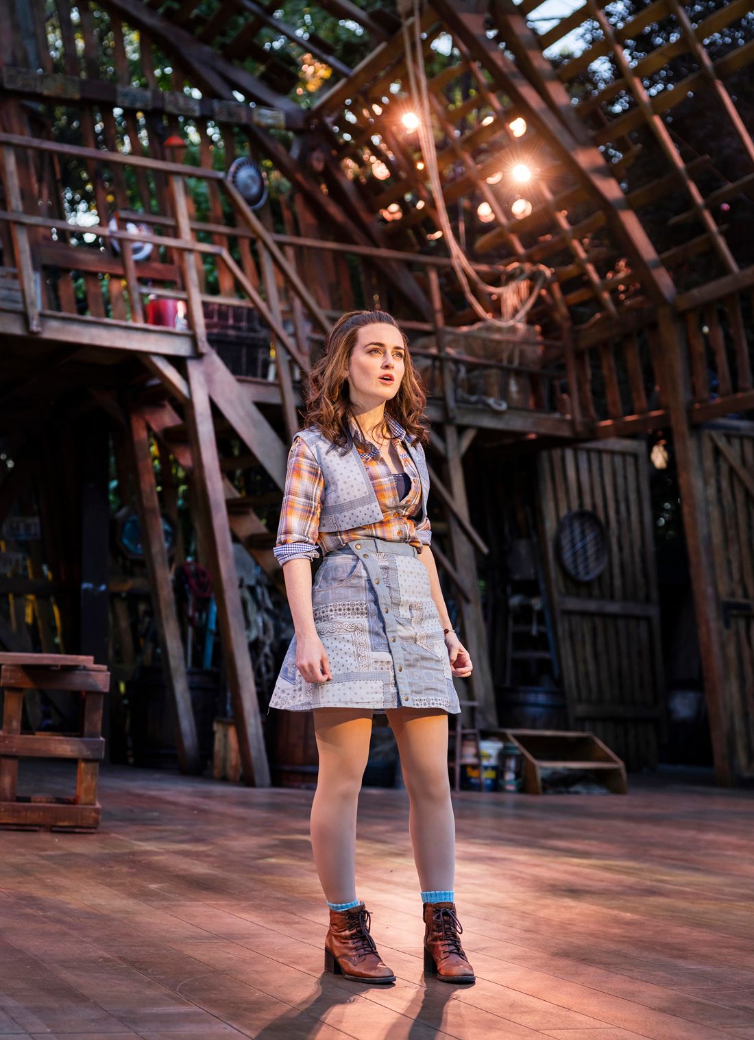 Sophie McShera as Maizy stands centre stage in a patchwork denim dress and checked shirt, mid-song, with warm lights glowing behind her.