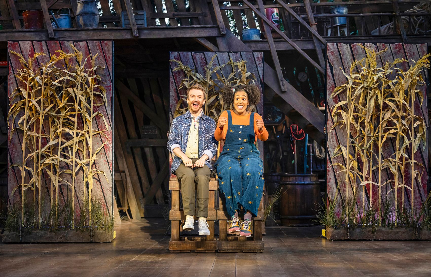 Steven Webb and Monique Ashe-Palmer as the storytellers sit side-by-side on wooden crates, miming driving with exaggerated, joyful expressions, flanked by corn-covered doors in a whimsical imaginary road trip moment.