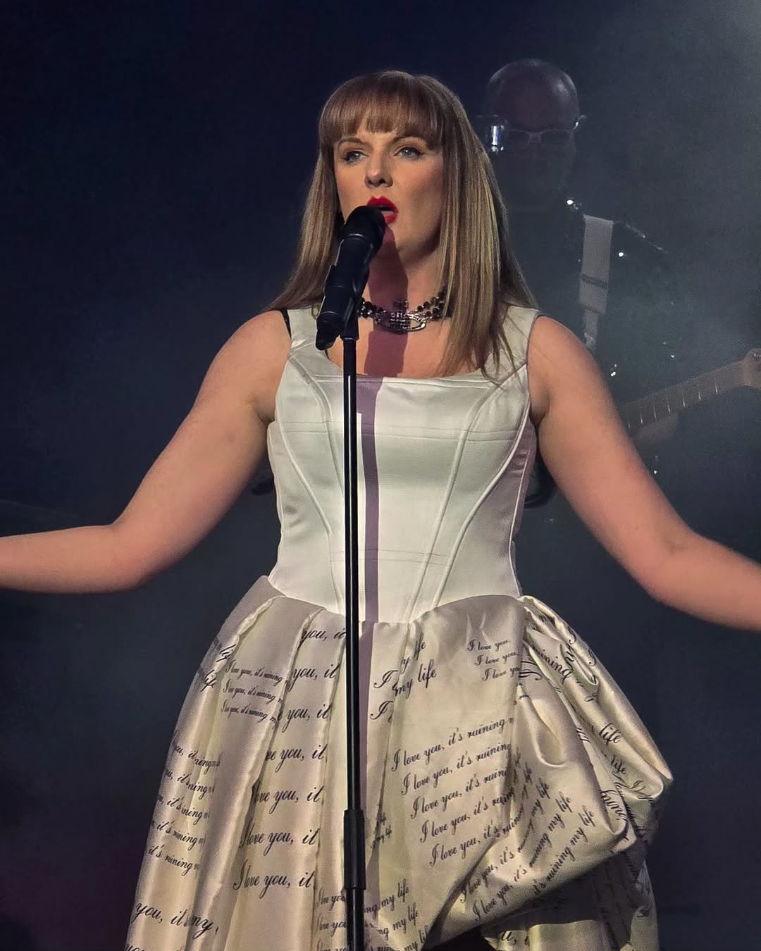 Close-up of Katy Ellis singing into a microphone wearing a white corset and cream puffball skirt printed with handwritten-style lyrics, styled after Taylor Swift’s Reputation or Lover eras.