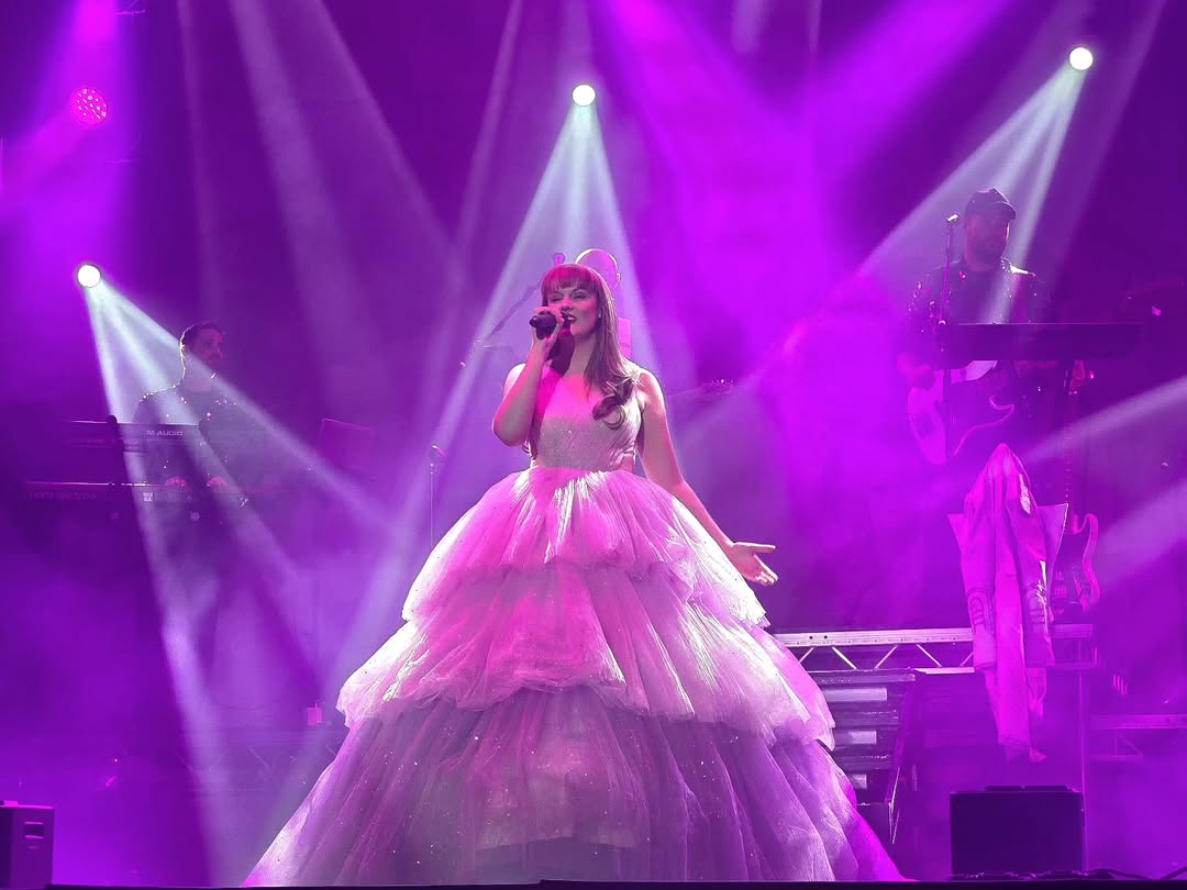 Katy Ellis performing in a voluminous pink fairy-tale gown surrounded by pink and purple stage lights, recreating the “Enchanted” look from Taylor Swift’s Speak Now era.