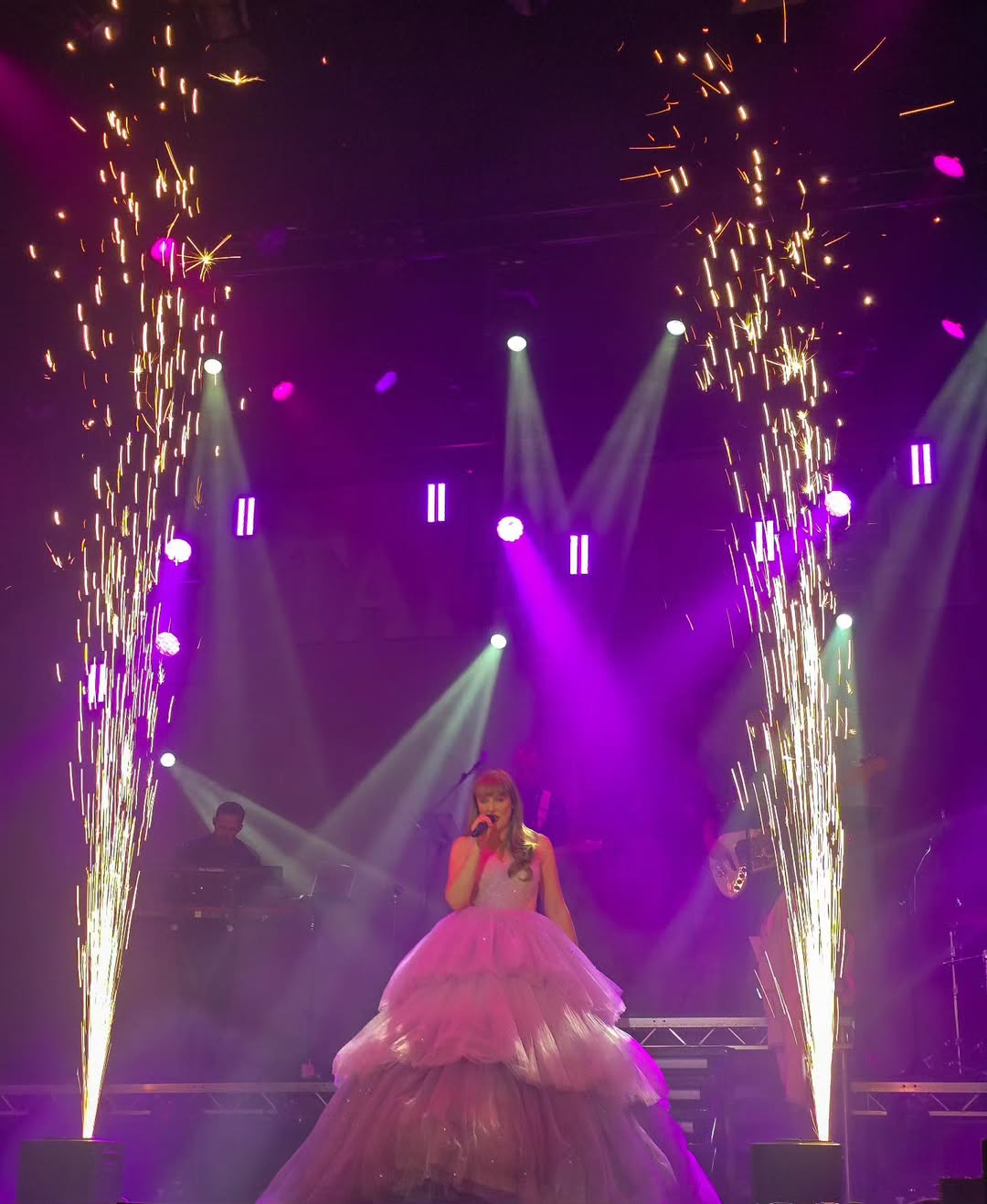 Katy Ellis in the same layered pink gown singing centre stage as golden sparks shoot up on either side, adding dramatic flair to the performance.