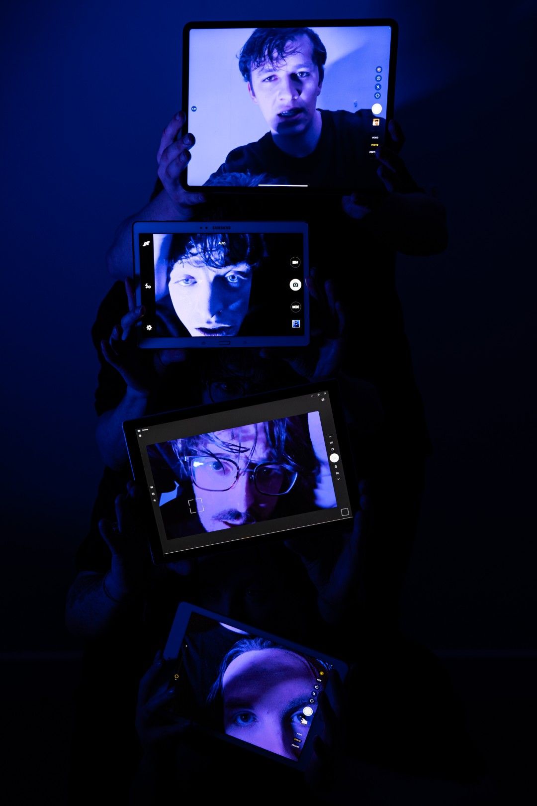 Four actors hold up tablets displaying intense close-up portraits of their faces, stacked vertically in a dark blue light, evoking a distorted video call.