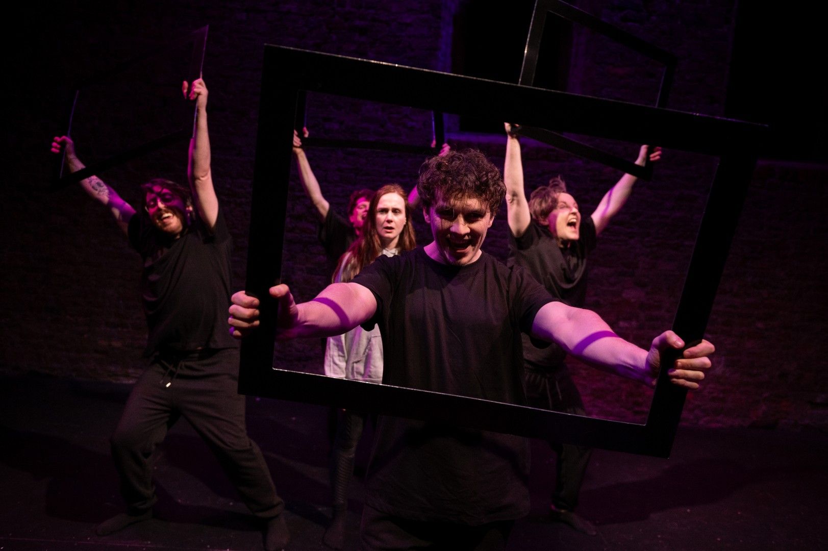 A man grins manically through a rectangular frame held toward the camera while other cast members behind him raise frames in a celebratory pose, bathed in magenta light.