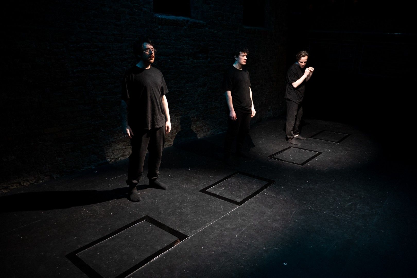 Three actors in black stand in isolated spotlight boxes on a bare stage, each framed by a black rectangle on the floor, creating a stark minimalist tableau.
