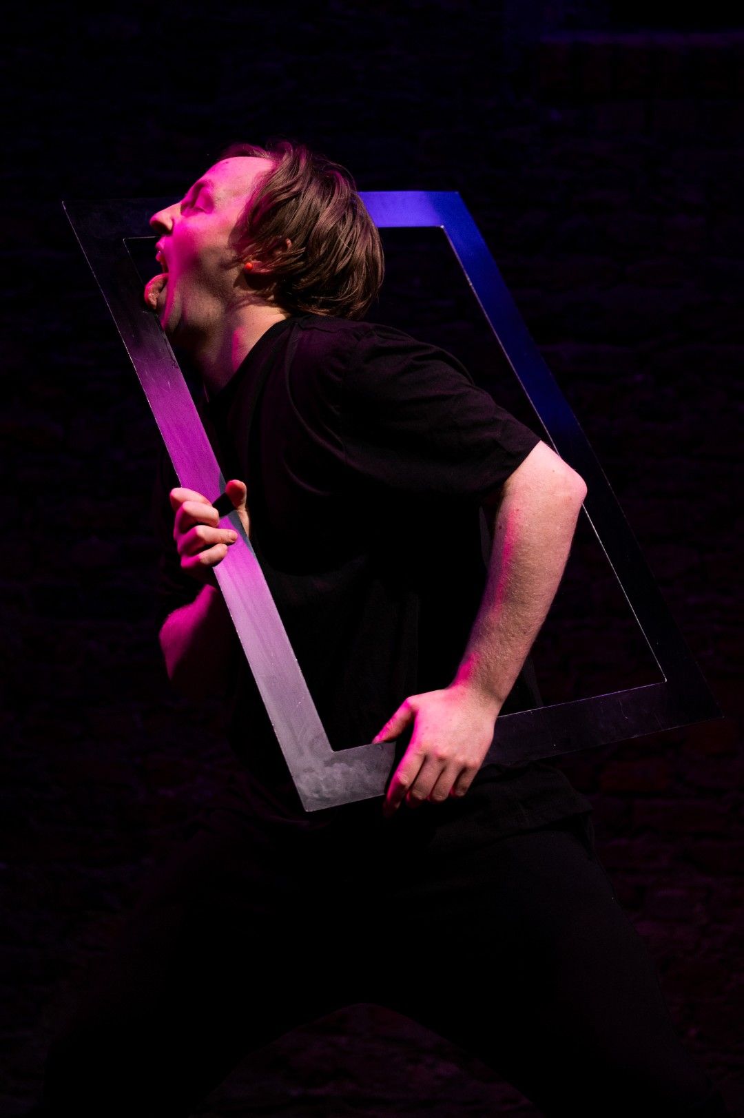 An actor sticks out his tongue dramatically while licking the inside edge of a rectangular frame he holds in front of his face, lit in theatrical purple and pink hues.