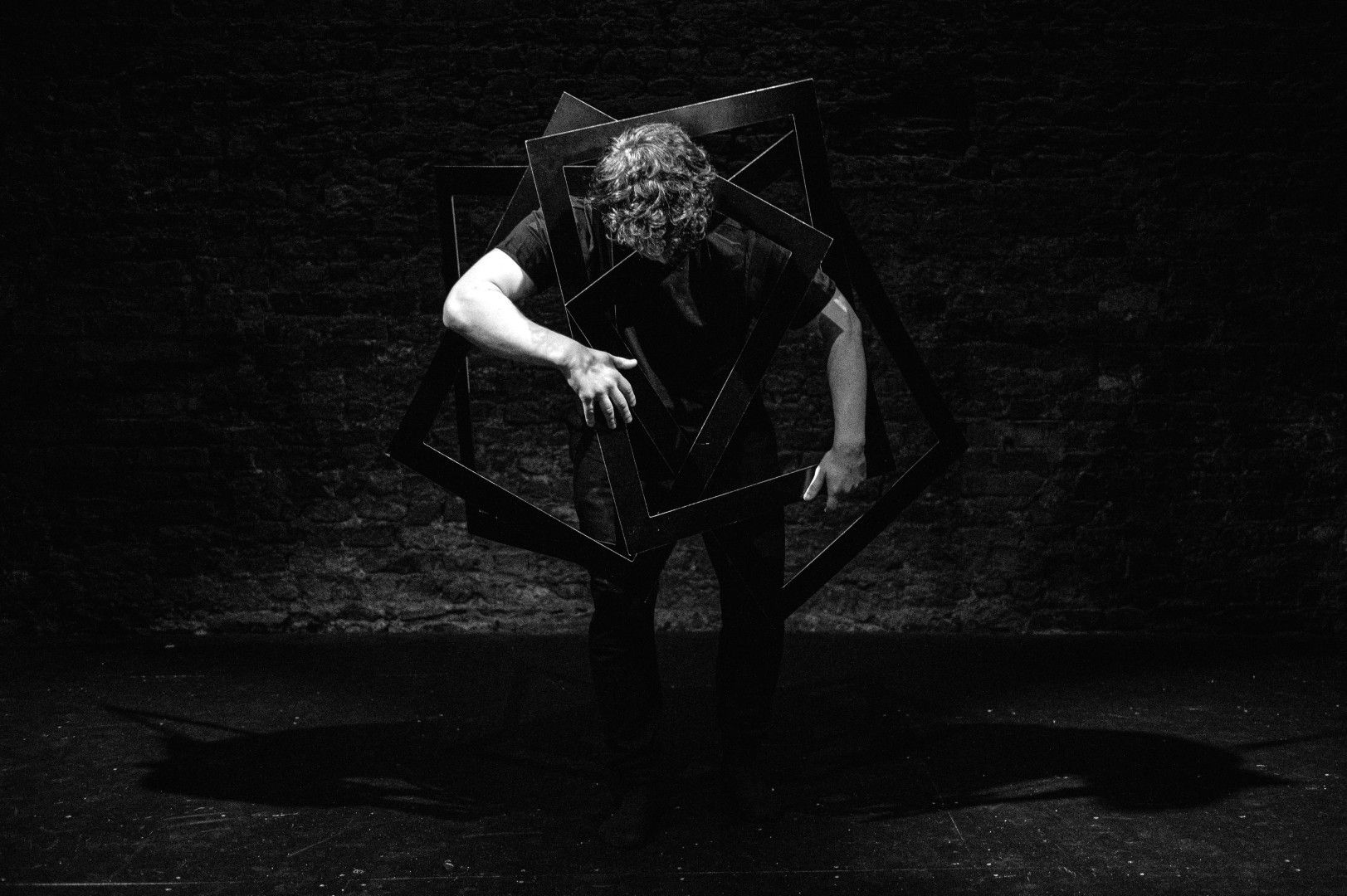 An actor hunches over inside a chaotic cluster of intersecting black frames, contorted in a pose that suggests entrapment or breakdown on a darkened stage.