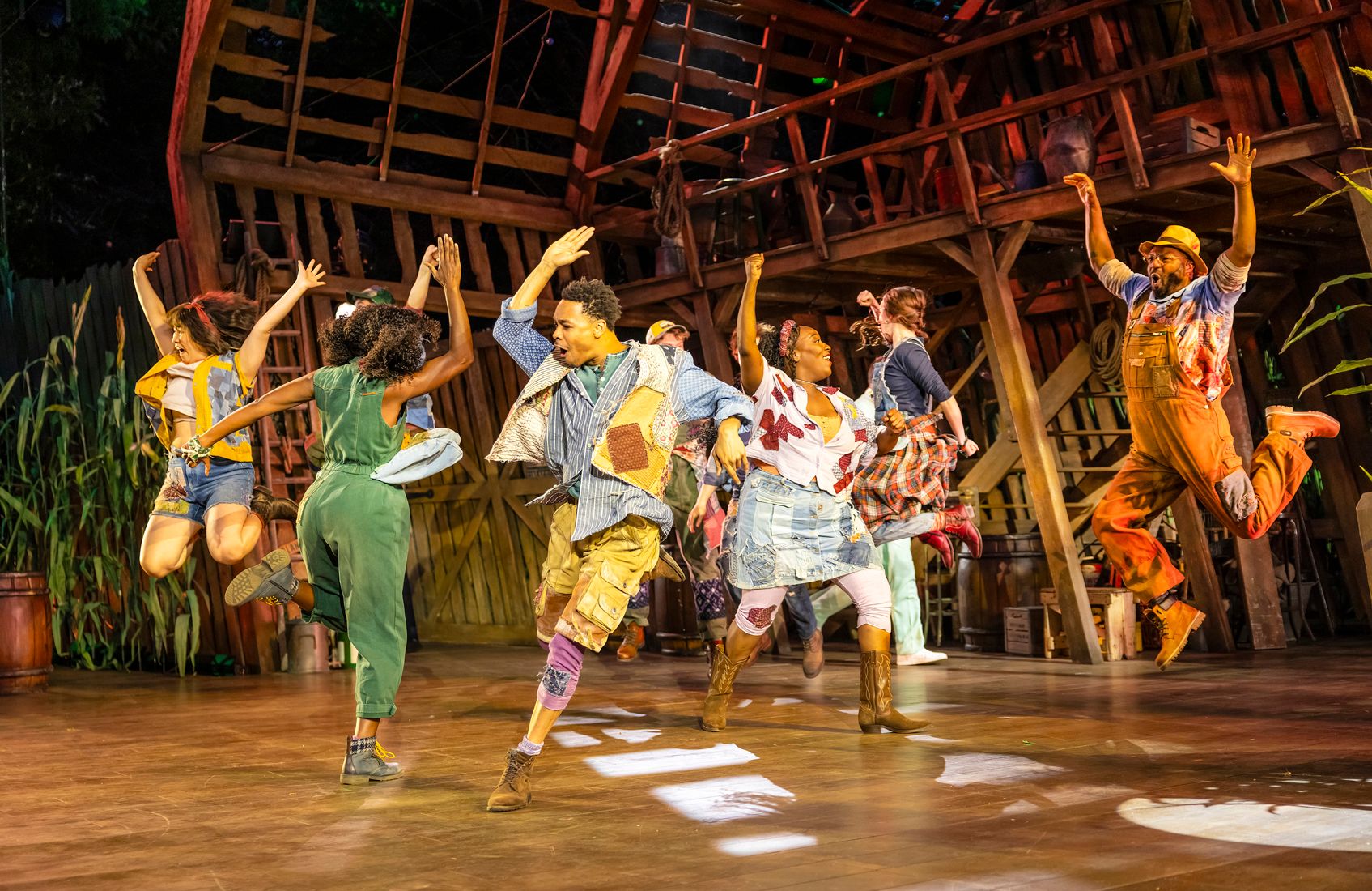 A high-energy moment mid-jump as cast members leap in all directions, dressed in denim, patches, and bright colours, with a vibrant barn set behind them.