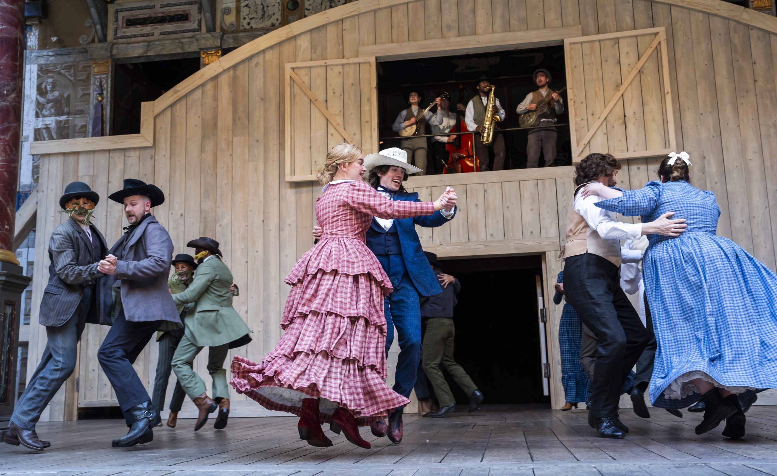 The company performs a lively group dance in front of the saloon doors, with Juliet in pink plaid and other characters mid-jump, while the band plays from the balcony.