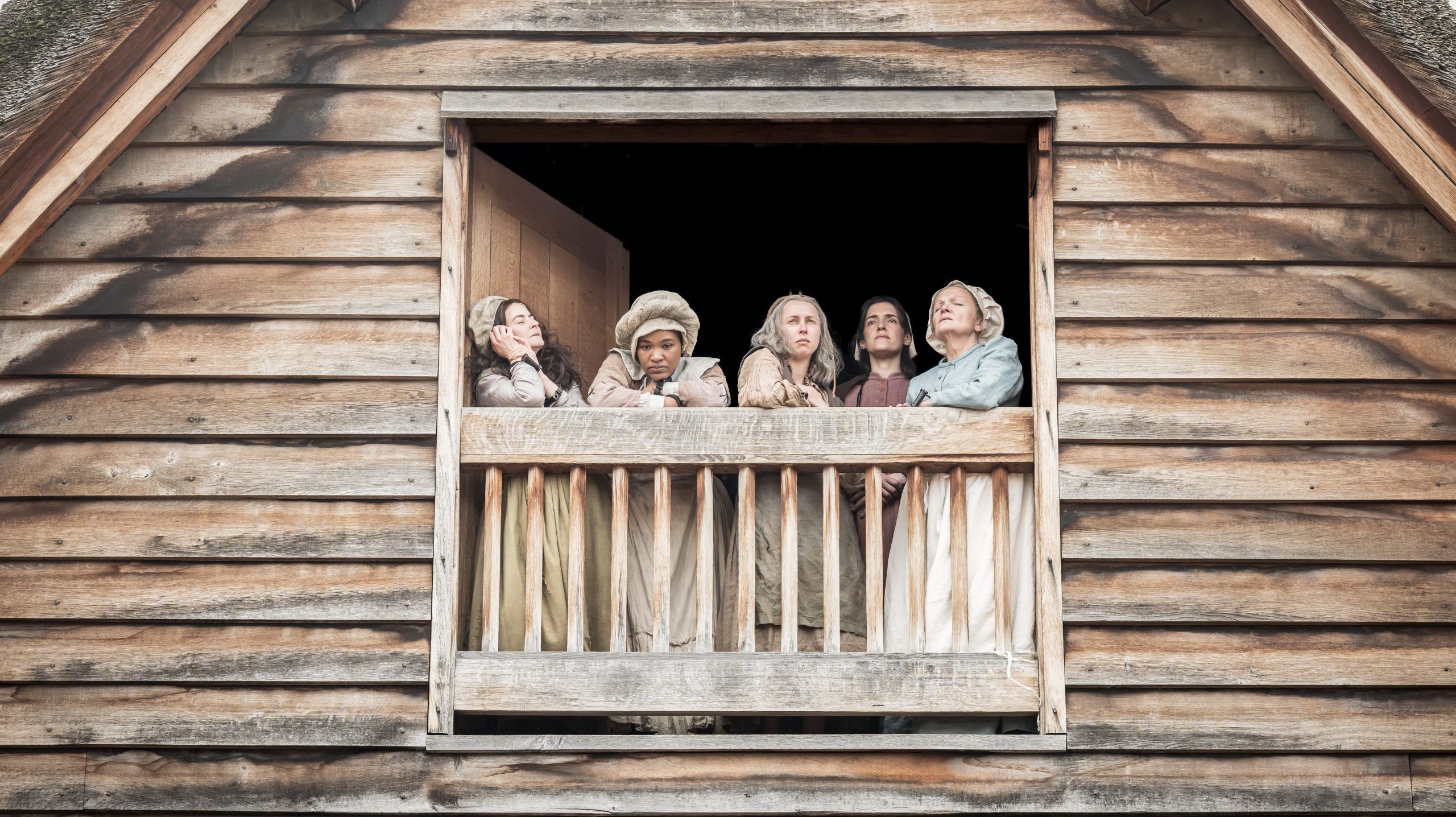 A row of women in bonnets and pale dresses lean out over the upper balcony of a timbered house set, each lost in thought — a moment of stillness before the storm.