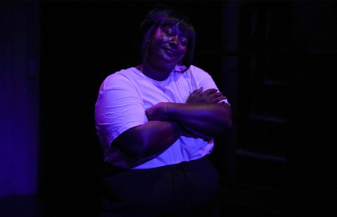 Yolanda Mercy stands under purple stage lighting with her arms crossed, smiling softly with her eyes closed.