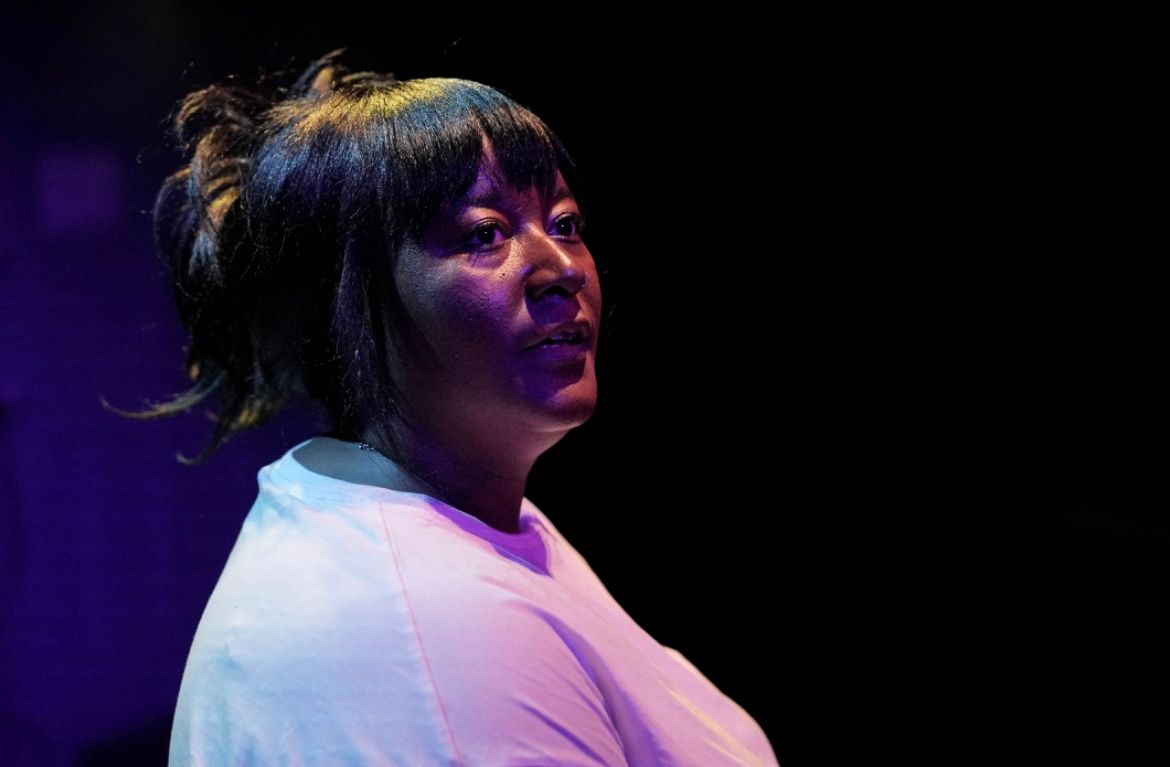 Yolanda Mercy stands under dramatic stage lighting in a white T-shirt, looking slightly off to the side with an attentive expression.
