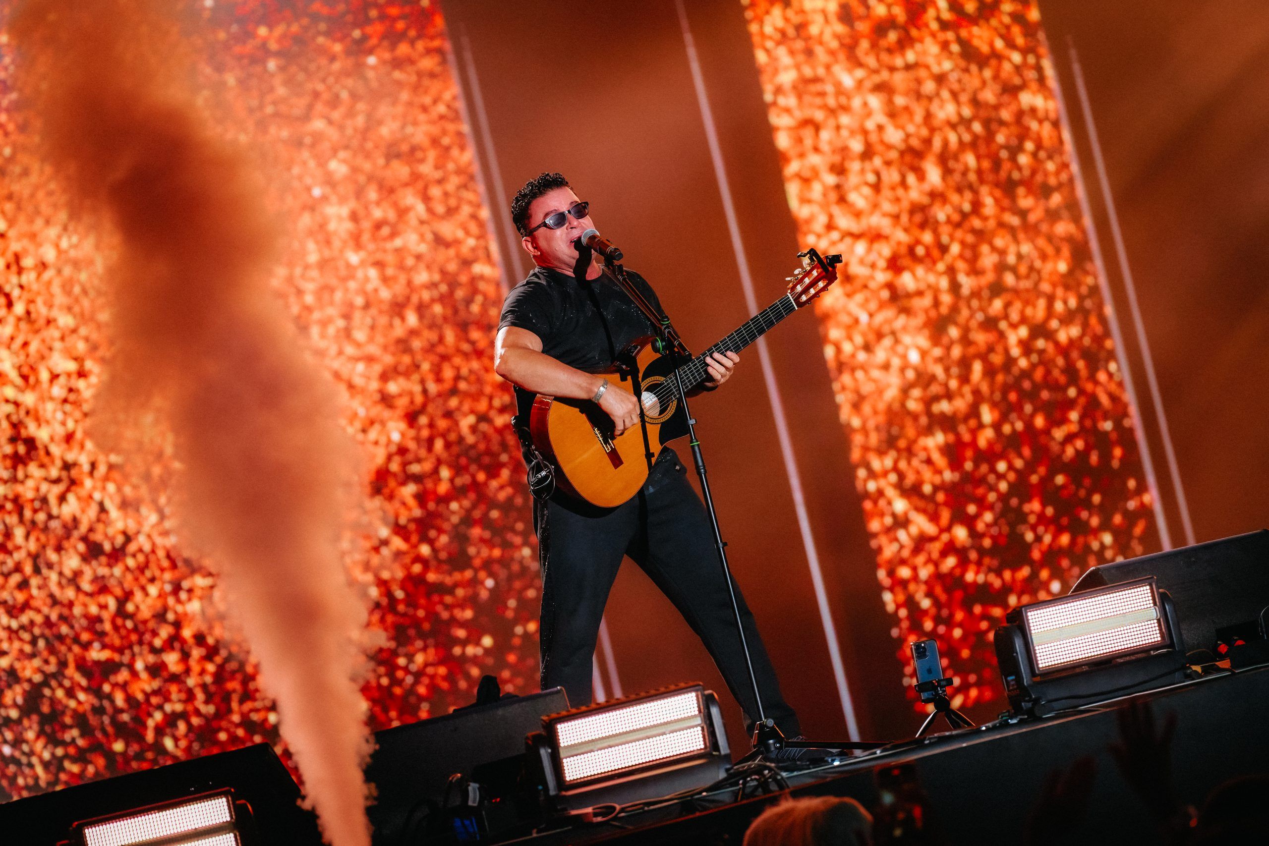 Male performer, No Mercy’s Marty Cintron, in a black outfit plays a guitar on a brightly lit stage at the Dream Fest concert. Smoke or fog rises from the side of the stage.